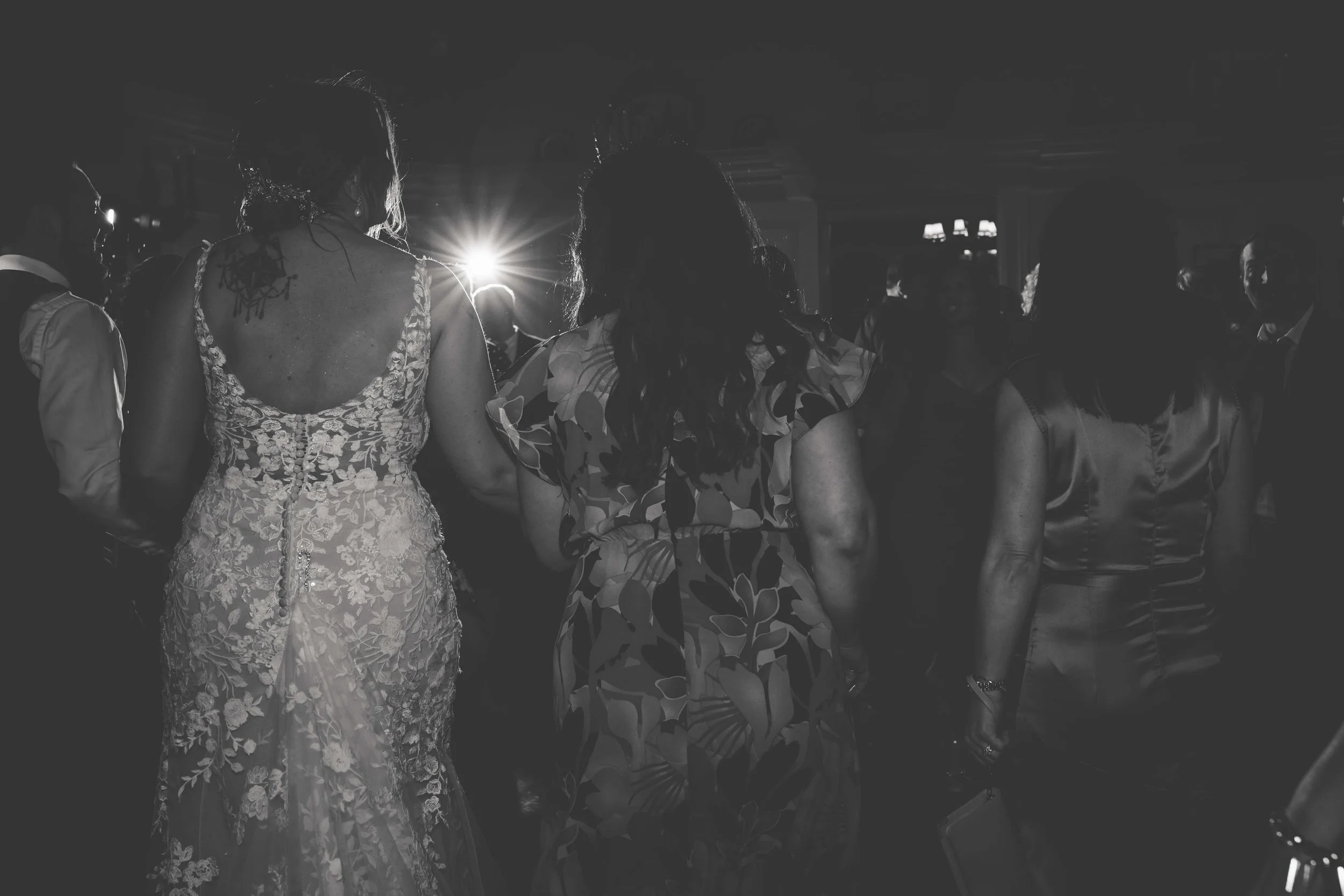 People dancing at a formal event, a woman in a lace wedding dress and others in elegant dresses, with a bright light behind them.