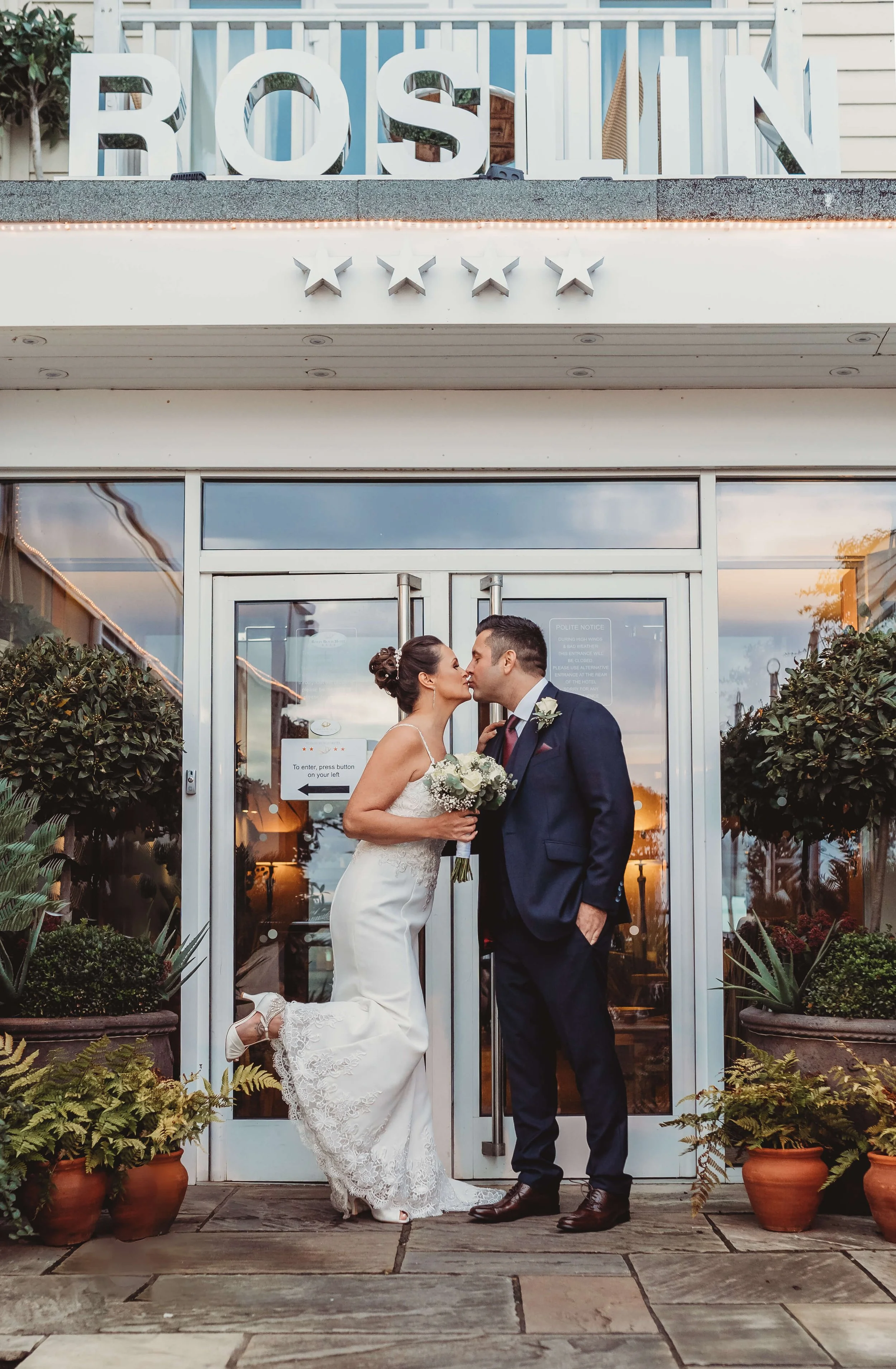 A bride and groom share a kiss outside a wedding venue decorated with potted plants, with the bride holding a bouquet, and the groom wearing a suit.