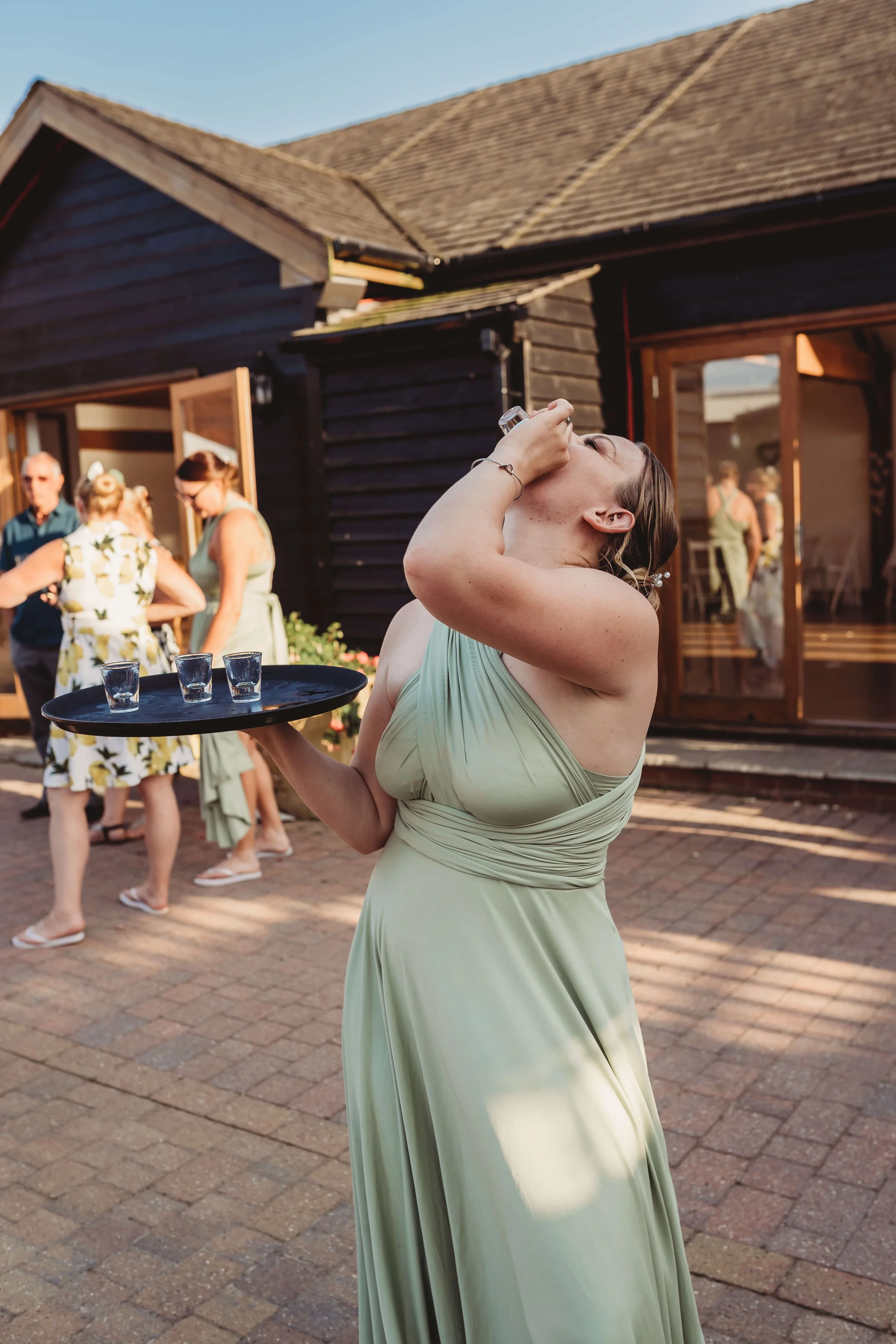 Woman in a light green dress tilting back and drinking from a shot glass while holding a tray with four shot glasses, at an outdoor gathering with others in the background.