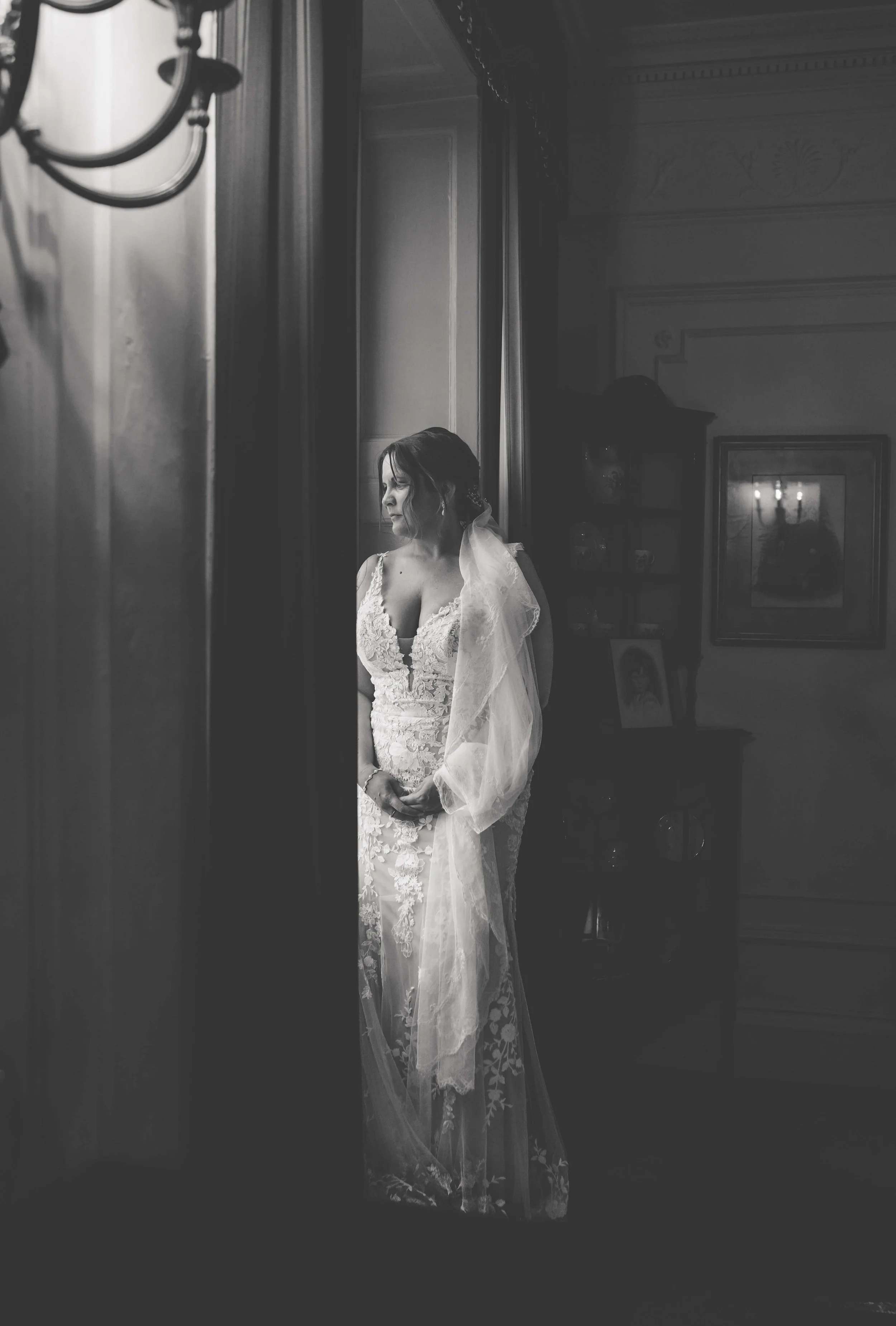 Black and white photo of a bride standing by a window in a lace wedding dress with a veil, looking contemplative.