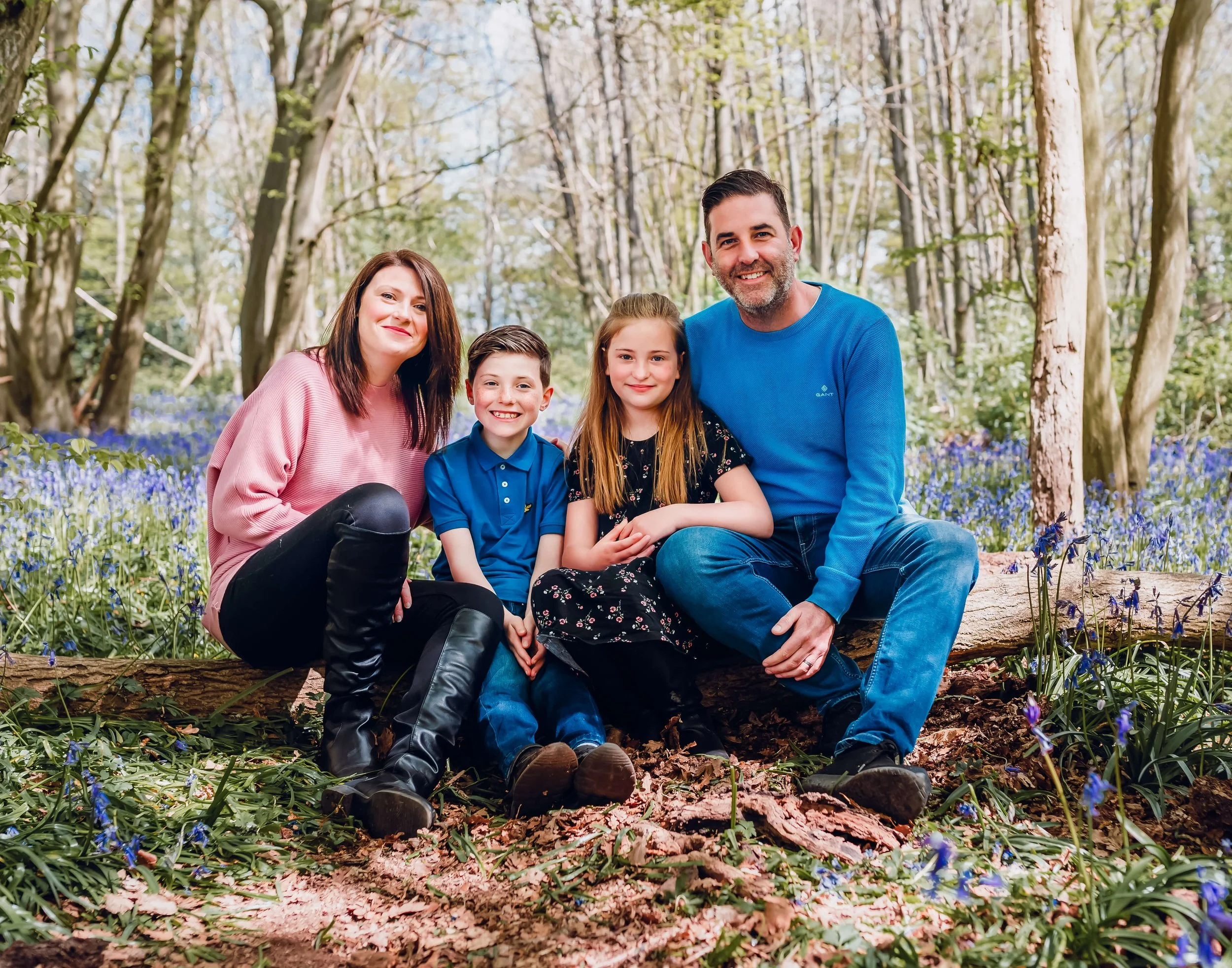 A family of four sitting on a fallen tree in a forest with blue flowers and tall trees surrounding them, smiling at the camera.