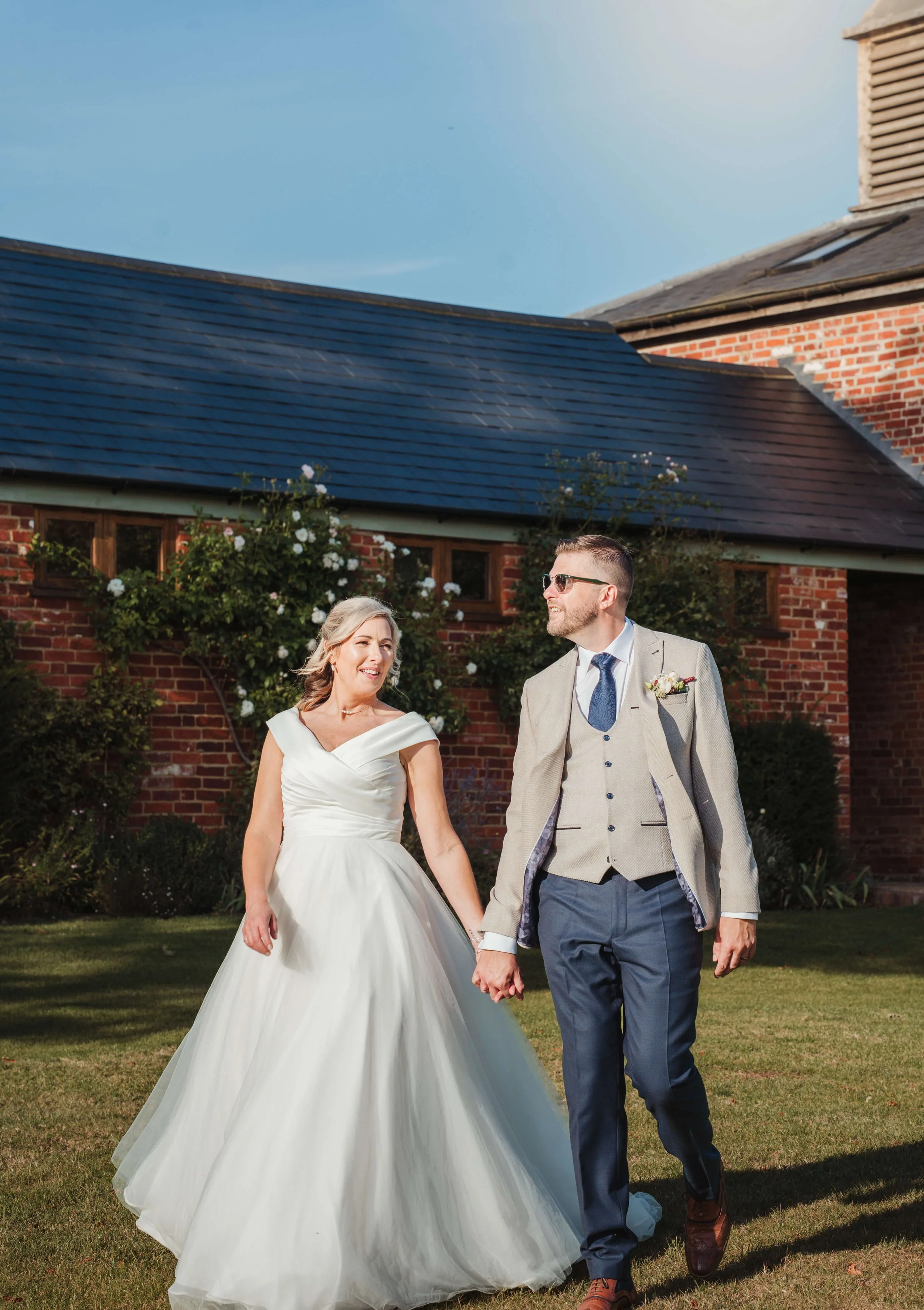 A bride and groom walking hand in hand on a grassy lawn during their wedding, with a brick building and blue sky in the background.