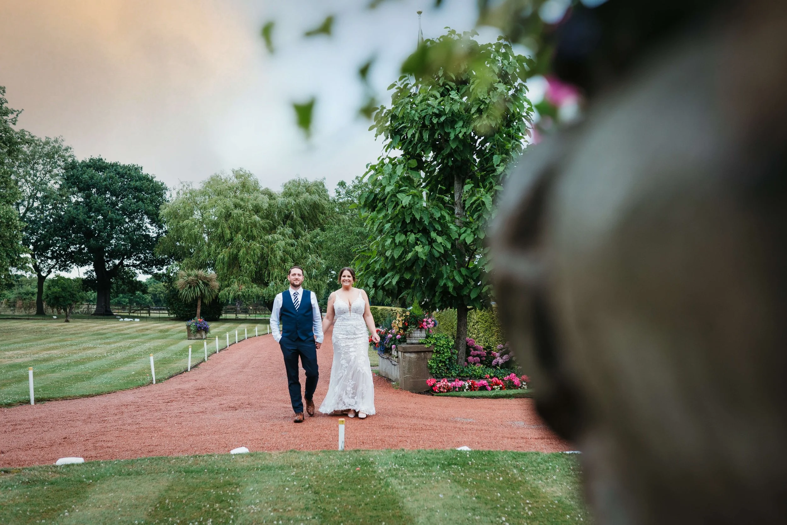 A bride and groom walking outdoors on a red gravel pathway surrounded by green trees and colorful flowers, with a large tree and cloudy sky in the background.