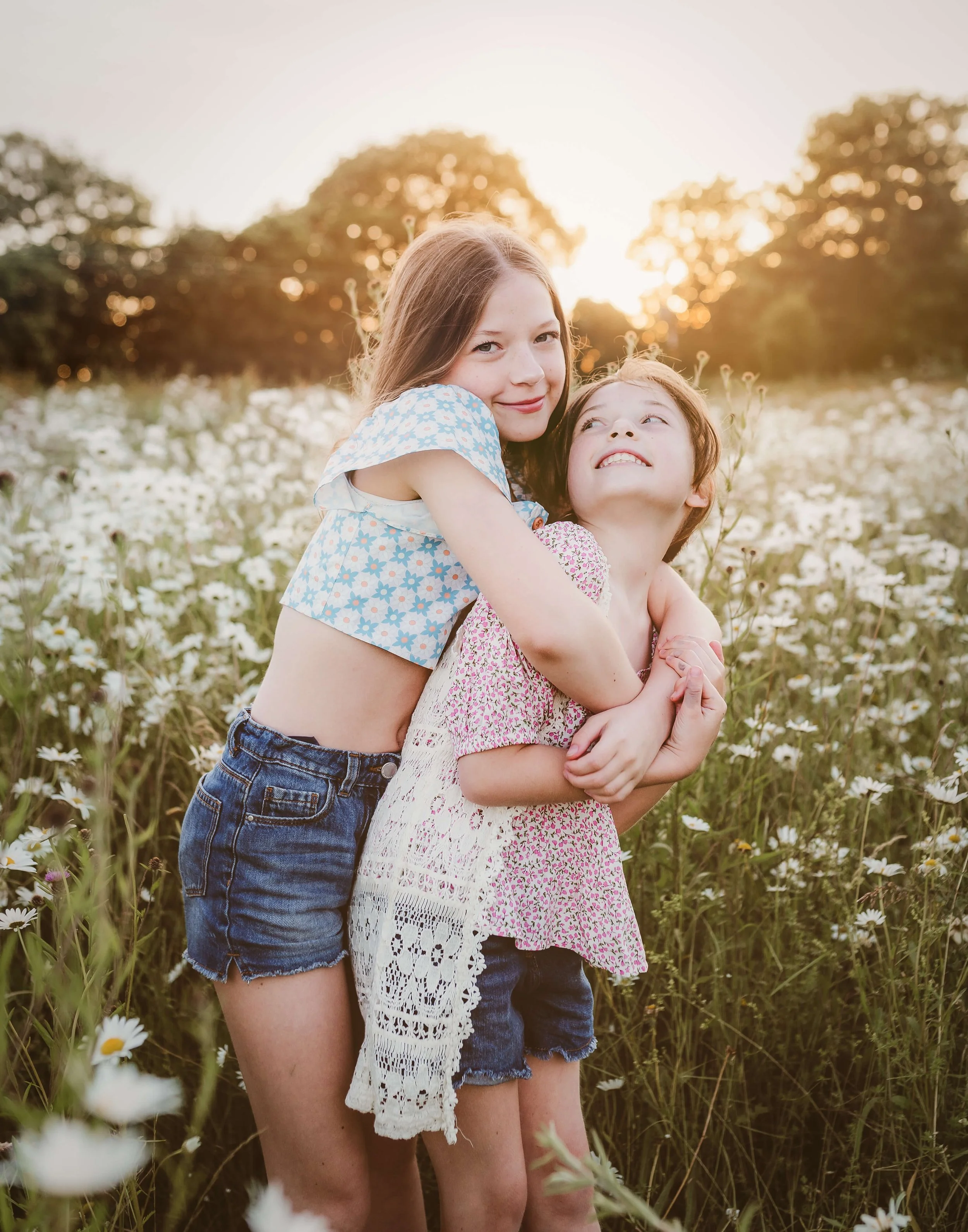 Two young girls hugging in a field of daisies at sunset, one with long brown hair and the other with short brown hair, both smiling.