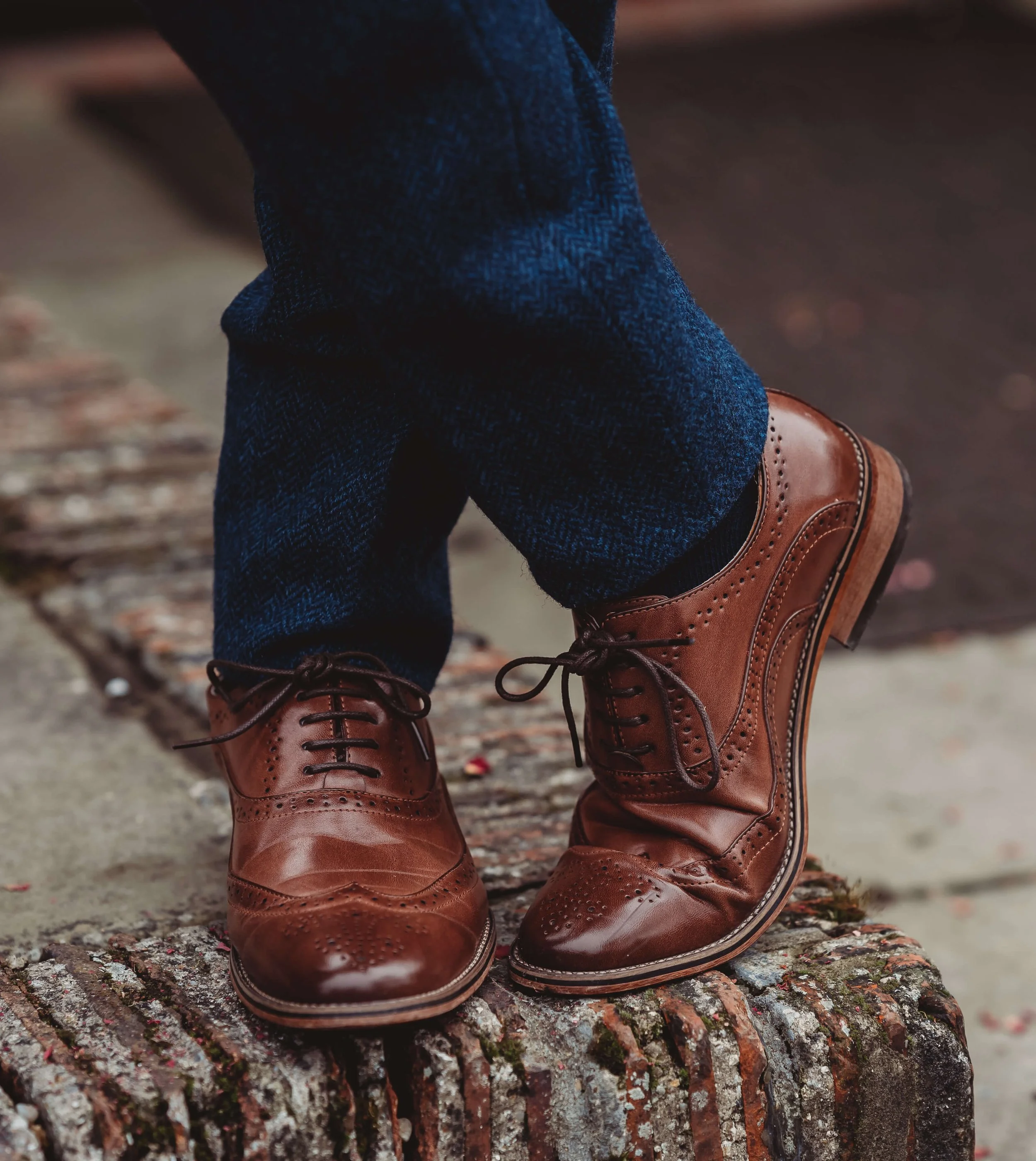 Close-up of a person wearing brown leather dress shoes and blue plaid trousers, standing on a weathered brick surface.