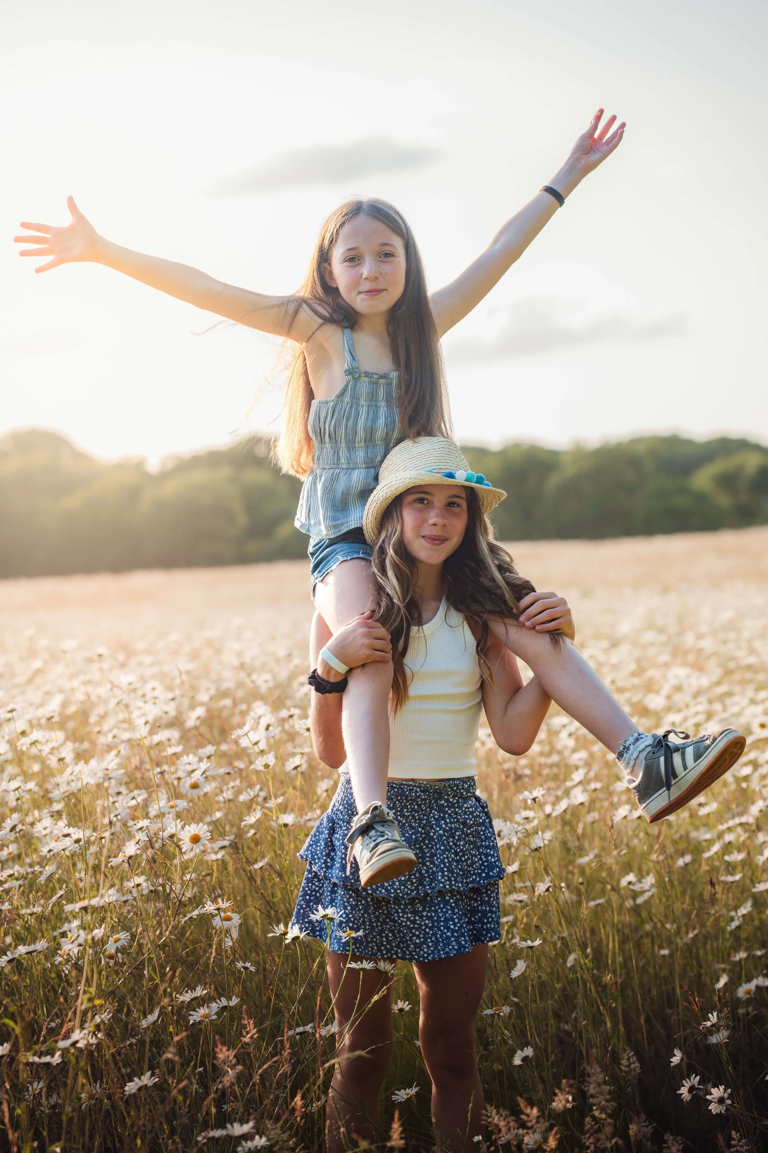 Two young girls in a field of daisies, one giving a piggyback ride on the other's shoulders, with arms raised in the air, during sunset.
