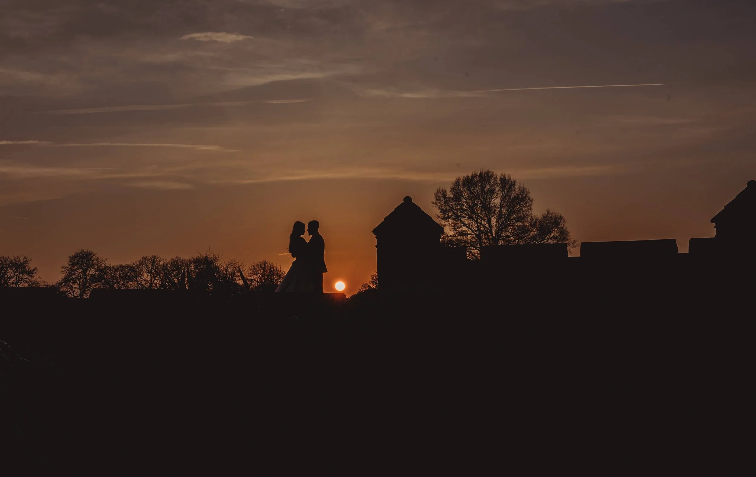 A silhouette of a couple standing close together on a hill at sunset with trees and buildings in the background.