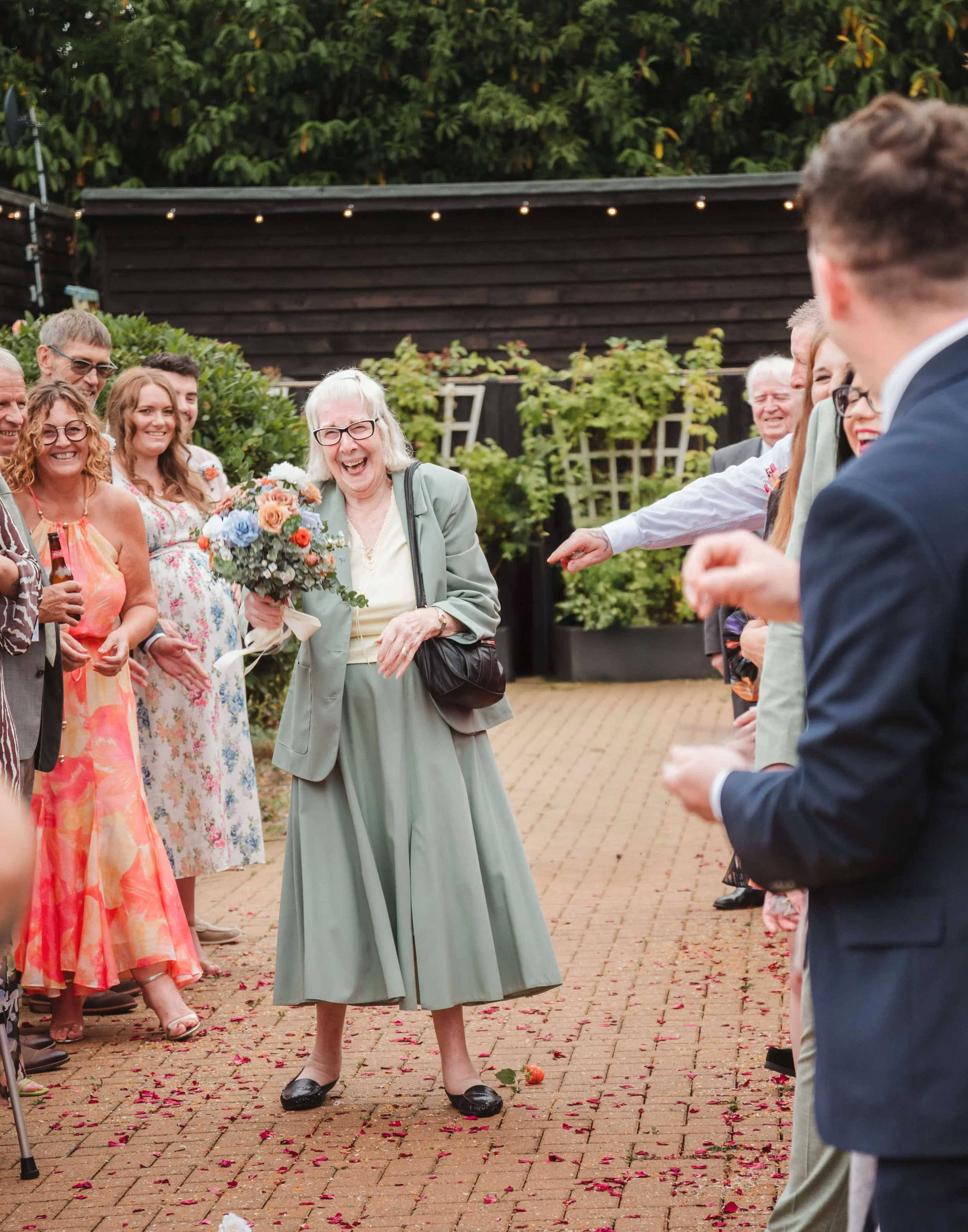 A group of people at a celebration, with an elderly woman in the center holding a bouquet of flowers, laughing among friends on a brick patio.