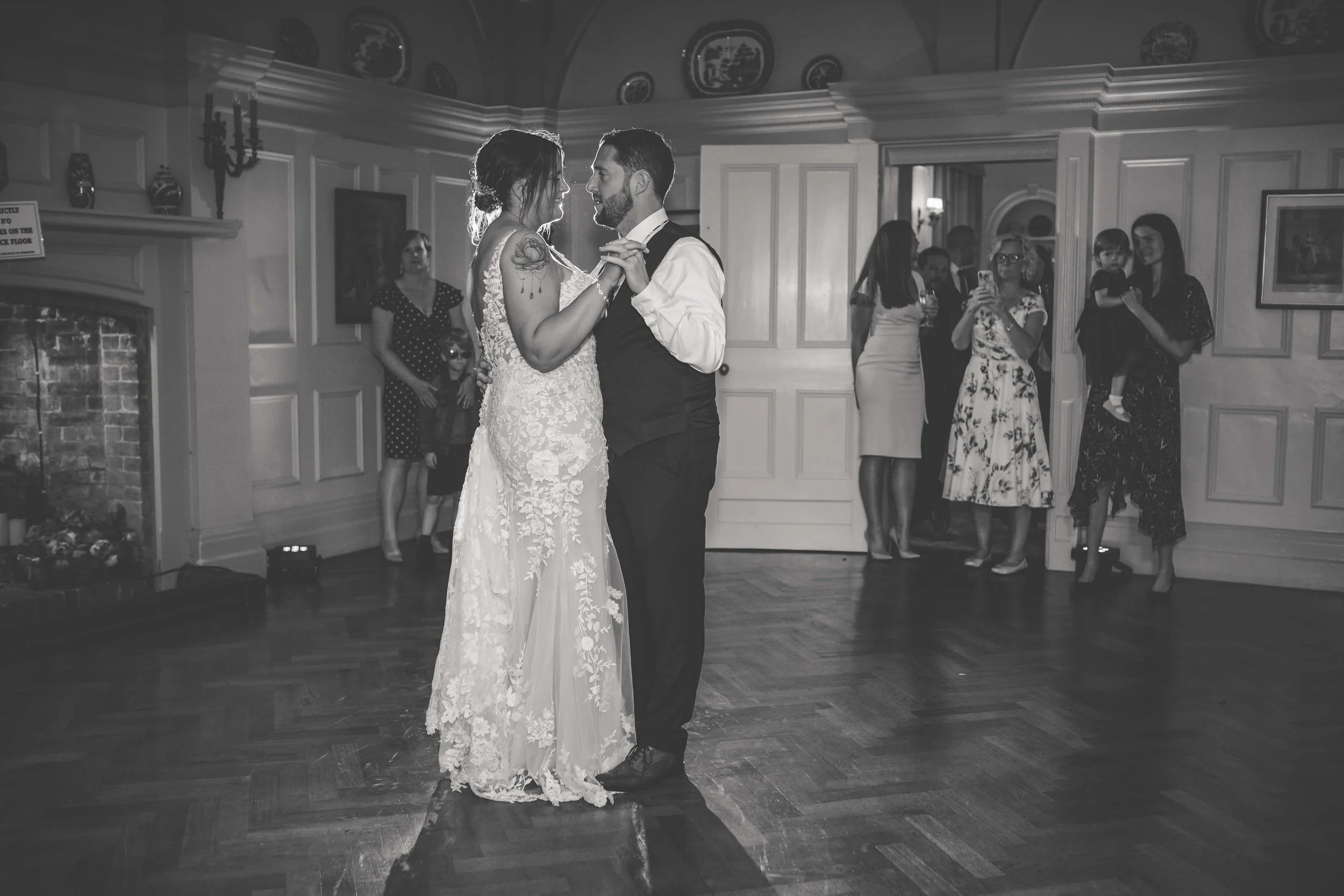 A couple is dancing during their wedding reception, surrounded by guests watching and taking photos, in an elegant ballroom with wooden floors.
