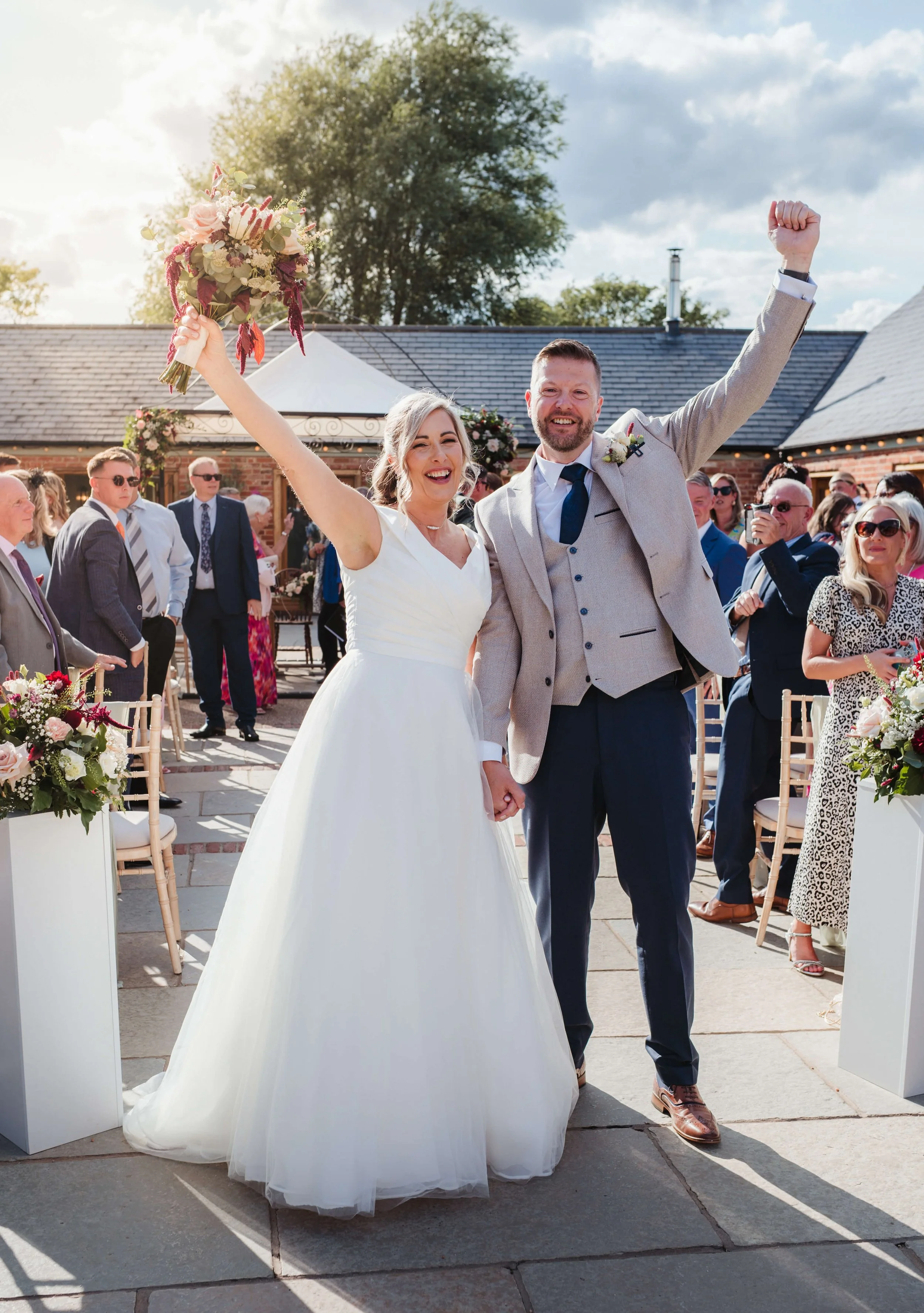 A newly married couple celebrating their wedding ceremony outdoors. The bride in a white wedding dress holding a bouquet, and the groom wearing a gray suit with a blue tie, are holding hands and smiling, with the bride raising her bouquet in celebrat