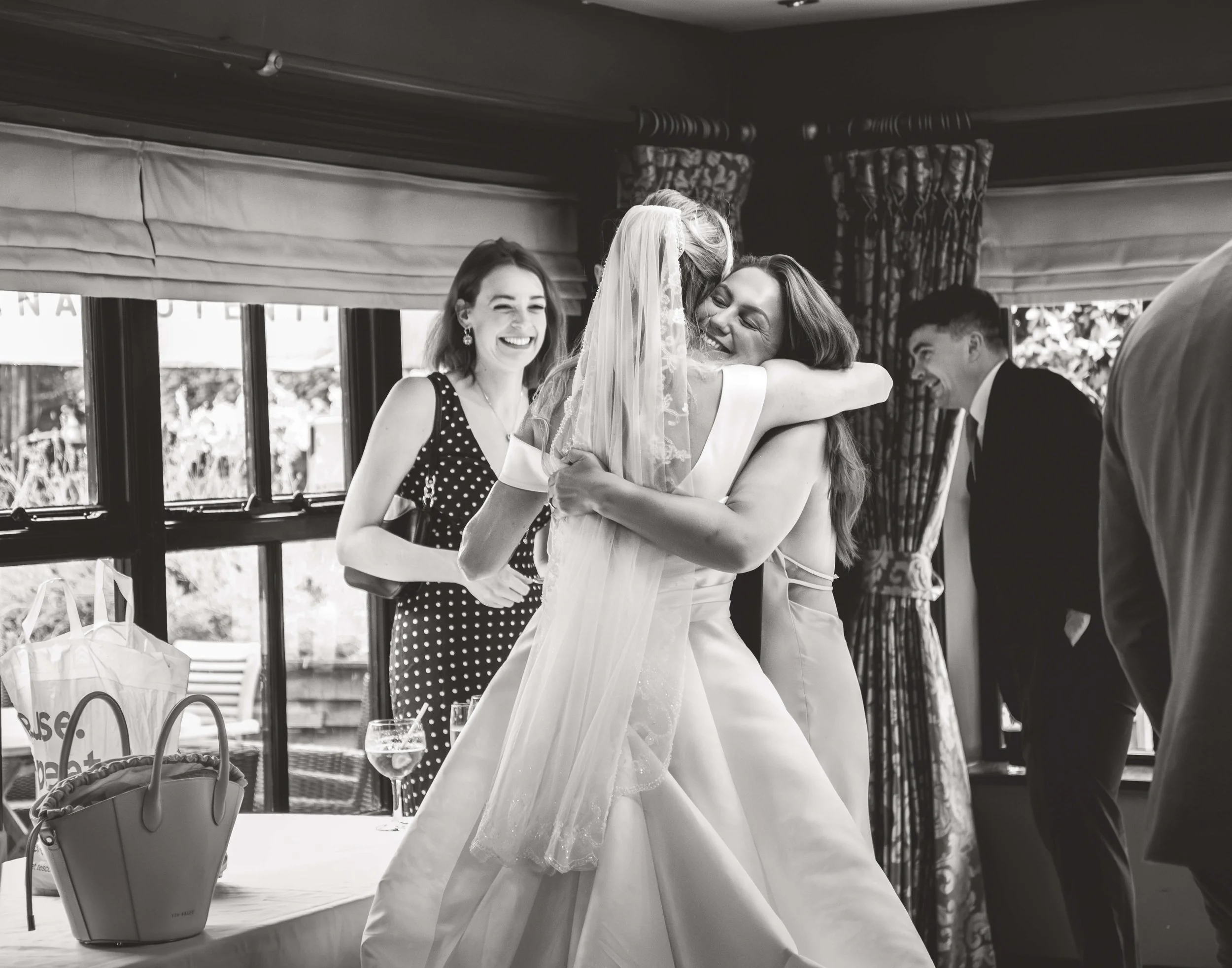 Two women embrace in a hug, one in a wedding dress and veil, the other in a sleeveless dress, smiling and happy. Two other people, a woman in a polka dot dress and a man in a suit, are nearby smiling. The scene is indoors with large windows and curta