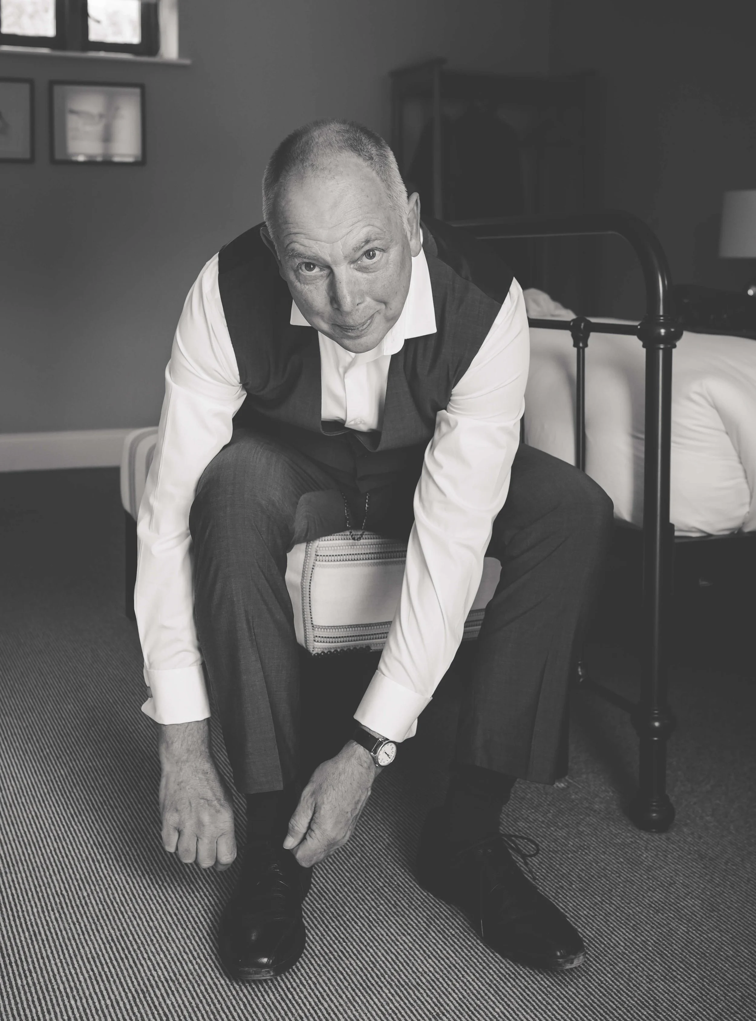 A man in formal attire, wearing a white shirt and dark vest, sitting on a small stool in a bedroom, leaning forward and tying the laces of his black dress shoes.