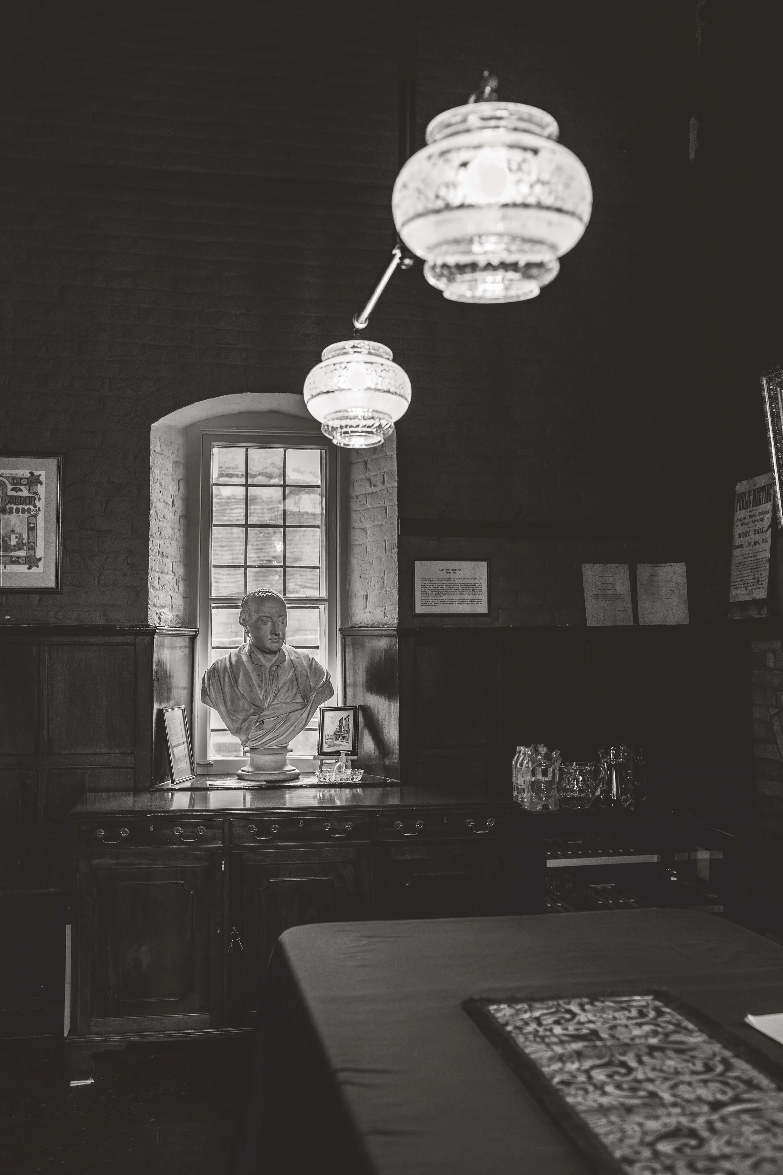 Black and white photo of an interior room with a bust sculpture of a man, a window behind it, and hanging ceiling lights.