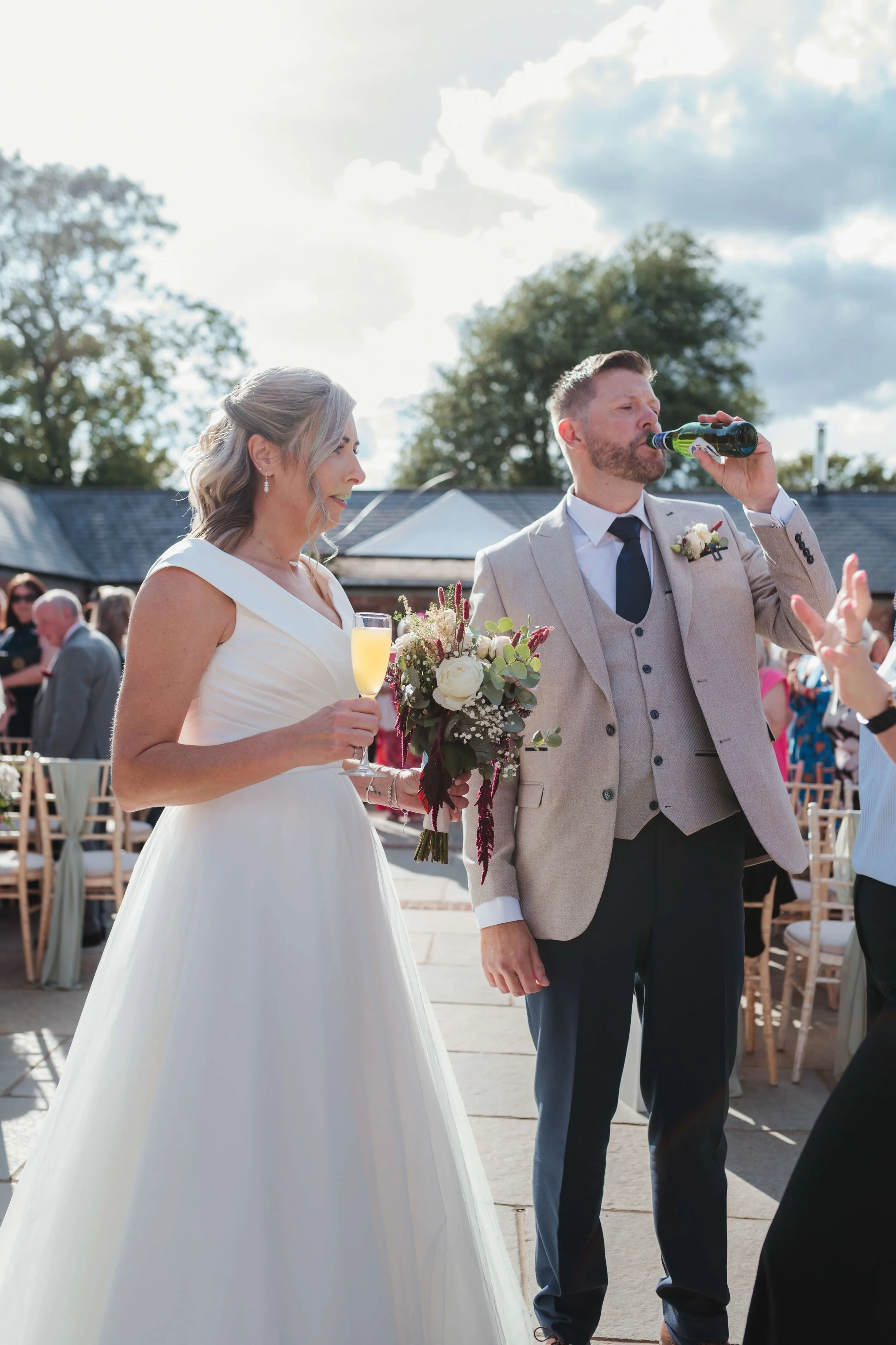 A bride in a white wedding dress holding a glass of champagne, standing next to a groom in a beige suit with a boutonniere, at an outdoor wedding reception. The groom is drinking from a bottle, and there are guests in the background under a partly cl