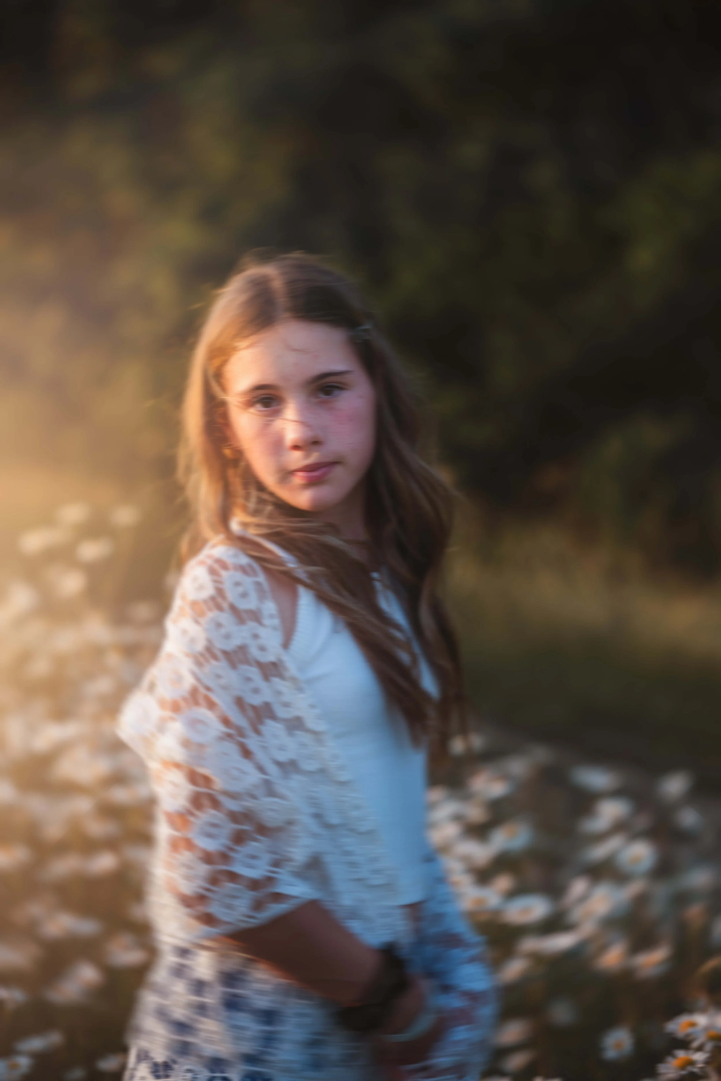 A young girl with long brown hair standing outdoors among flowers with a blurred background, wearing a white lace dress, during sunset.