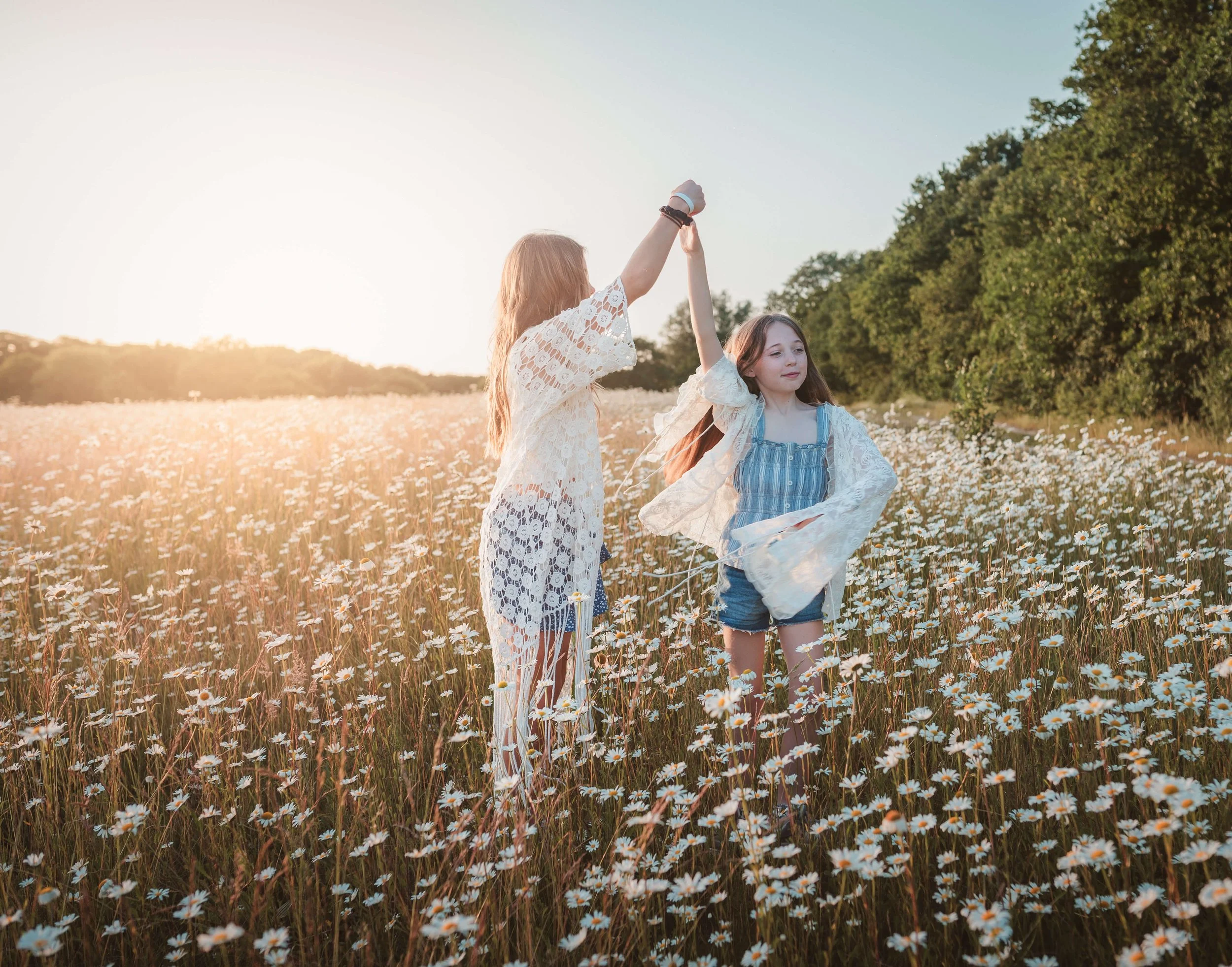 Two girls dancing hand in hand in a field of daisies during sunset.