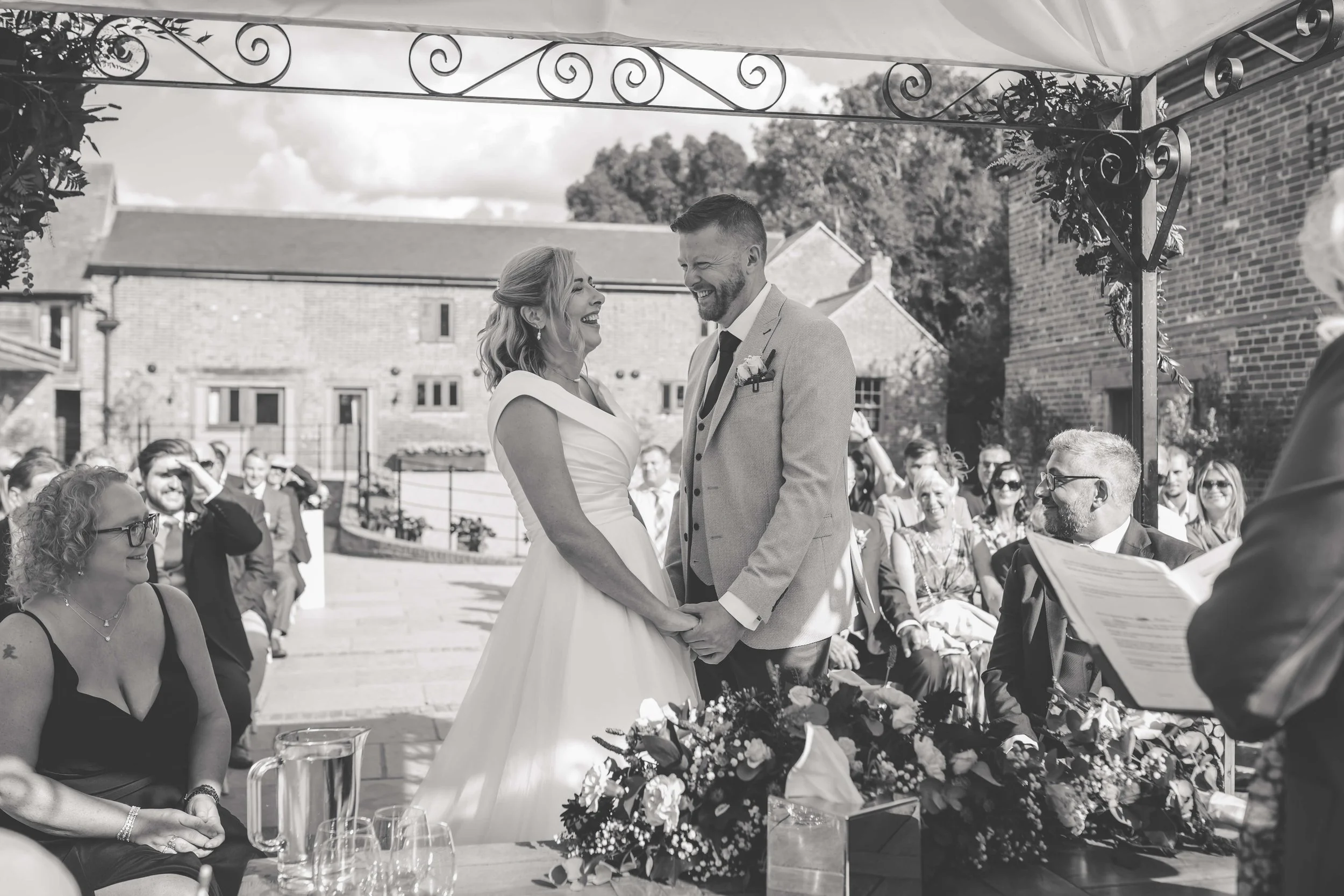 A black and white photo of a wedding ceremony outdoors with the bride and groom holding hands and smiling at each other under a canopy, surrounded by seated guests and onlookers, with a brick building in the background.