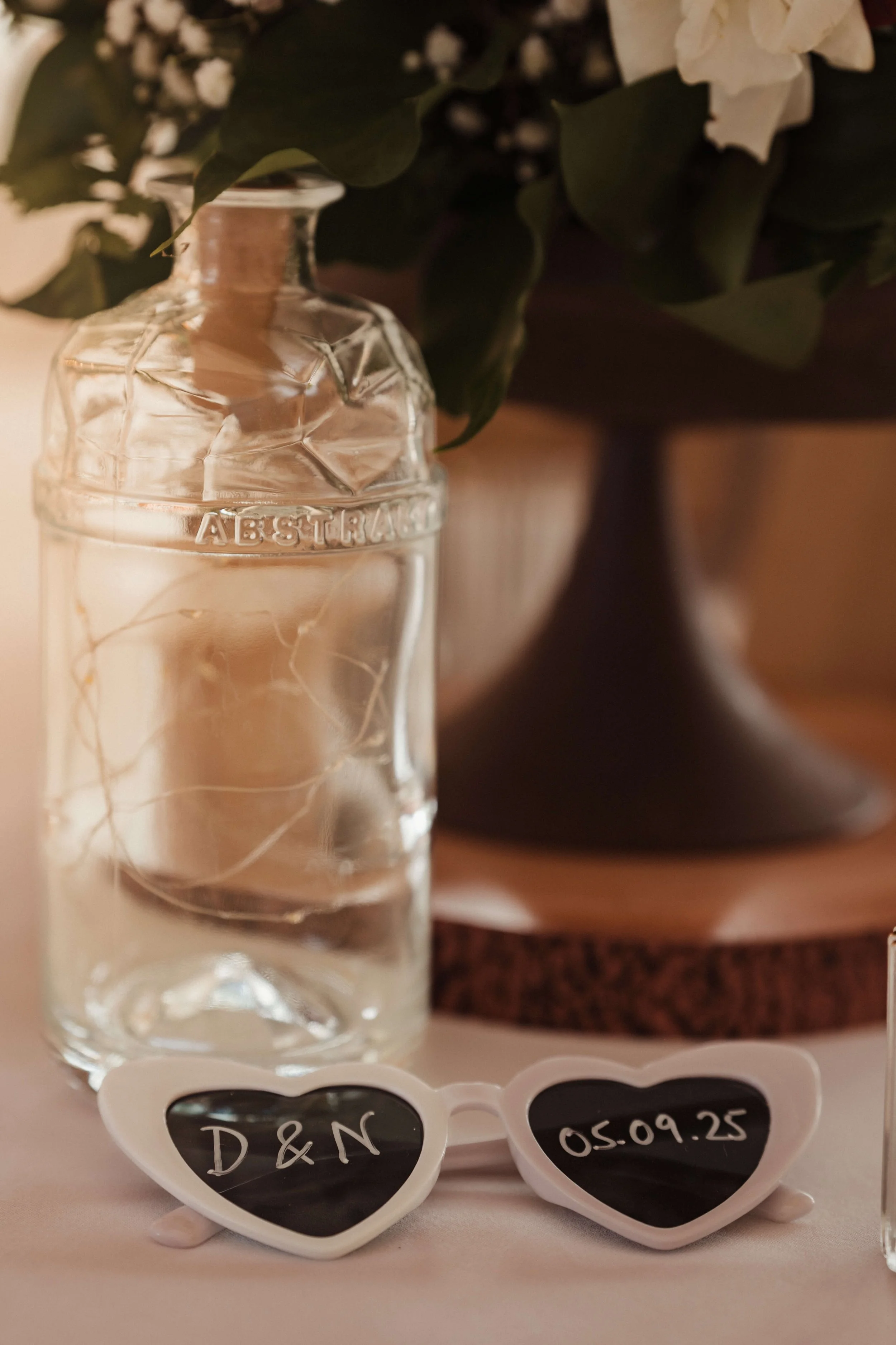 A glass bottle of clear liquid with roots inside, placed on a table with heart-shaped sunglasses display reading 'D & N' and date '05.09.25'. In the background, a flower arrangement with white flowers and green leaves.