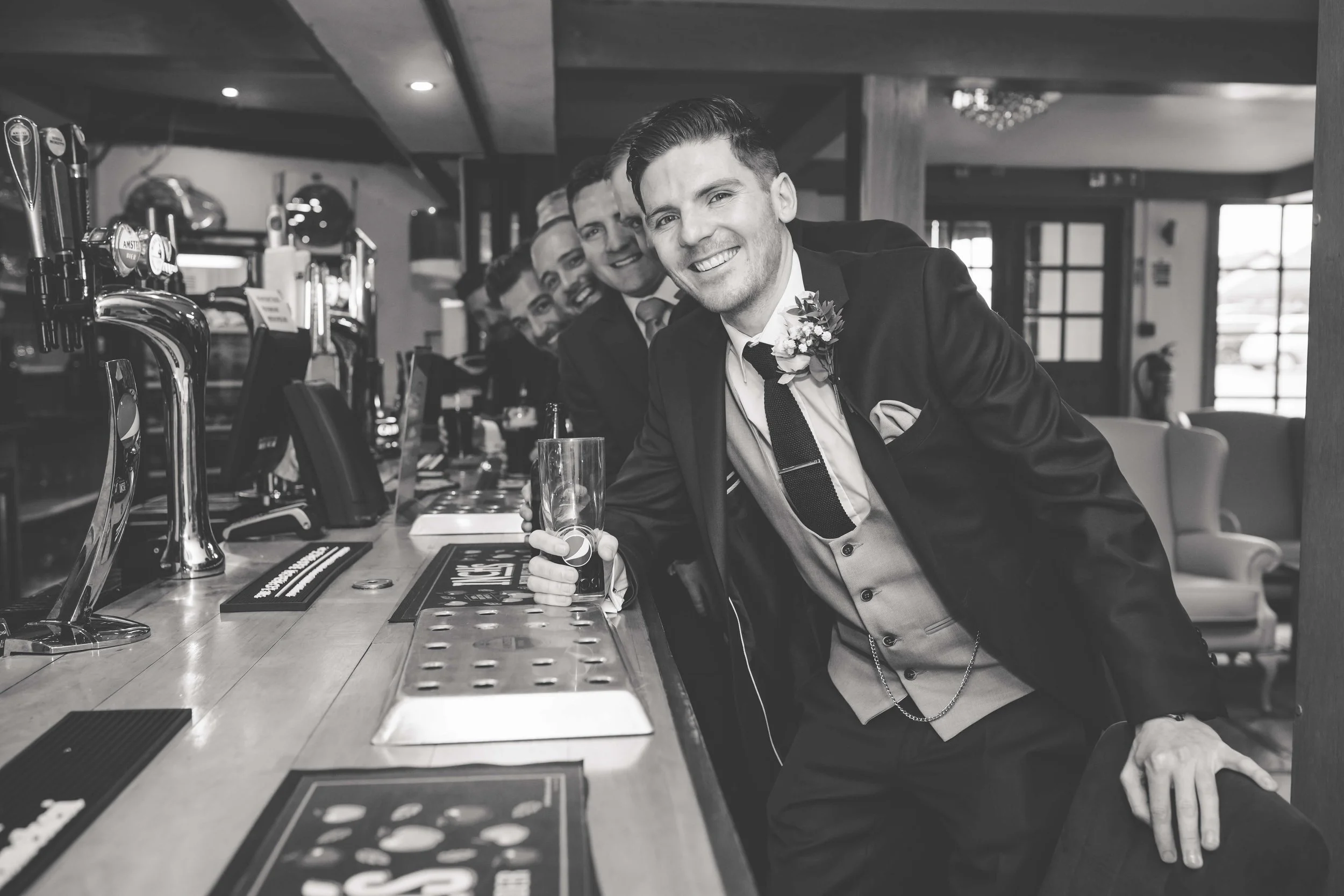 A group of smiling men in suits at a bar, with the man in front holding a glass, in a cozy, well-lit pub.
