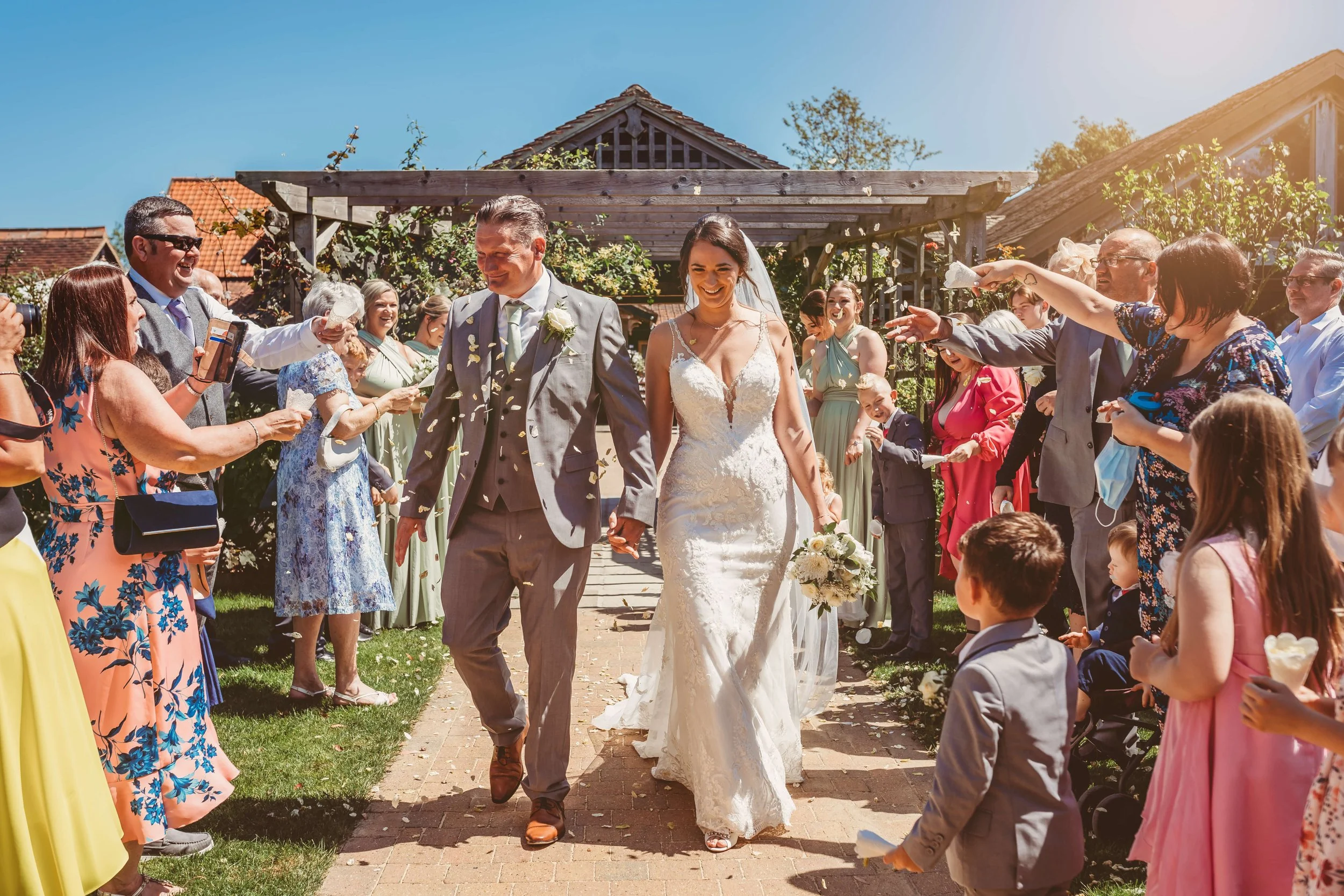 A bride and groom walking down the aisle after their wedding ceremony, surrounded by cheering guests throwing flower petals, outdoors in sunny weather.