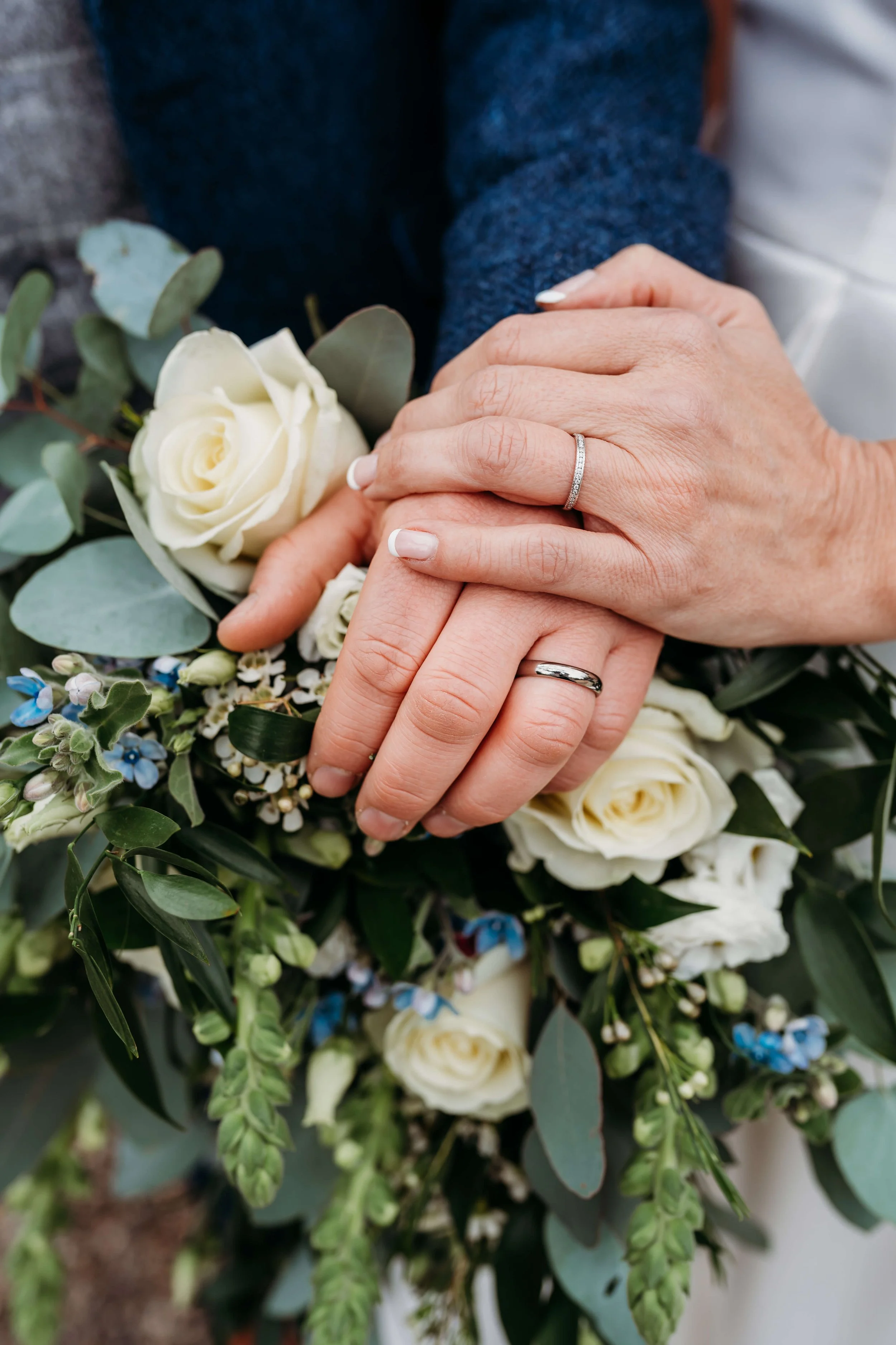 Close-up of a woman and man holding hands, showing wedding rings. The woman’s hand has a diamond wedding band, and the man's hand has a plain wedding band. The couple’s hands are resting on a bouquet of white roses and greenery.