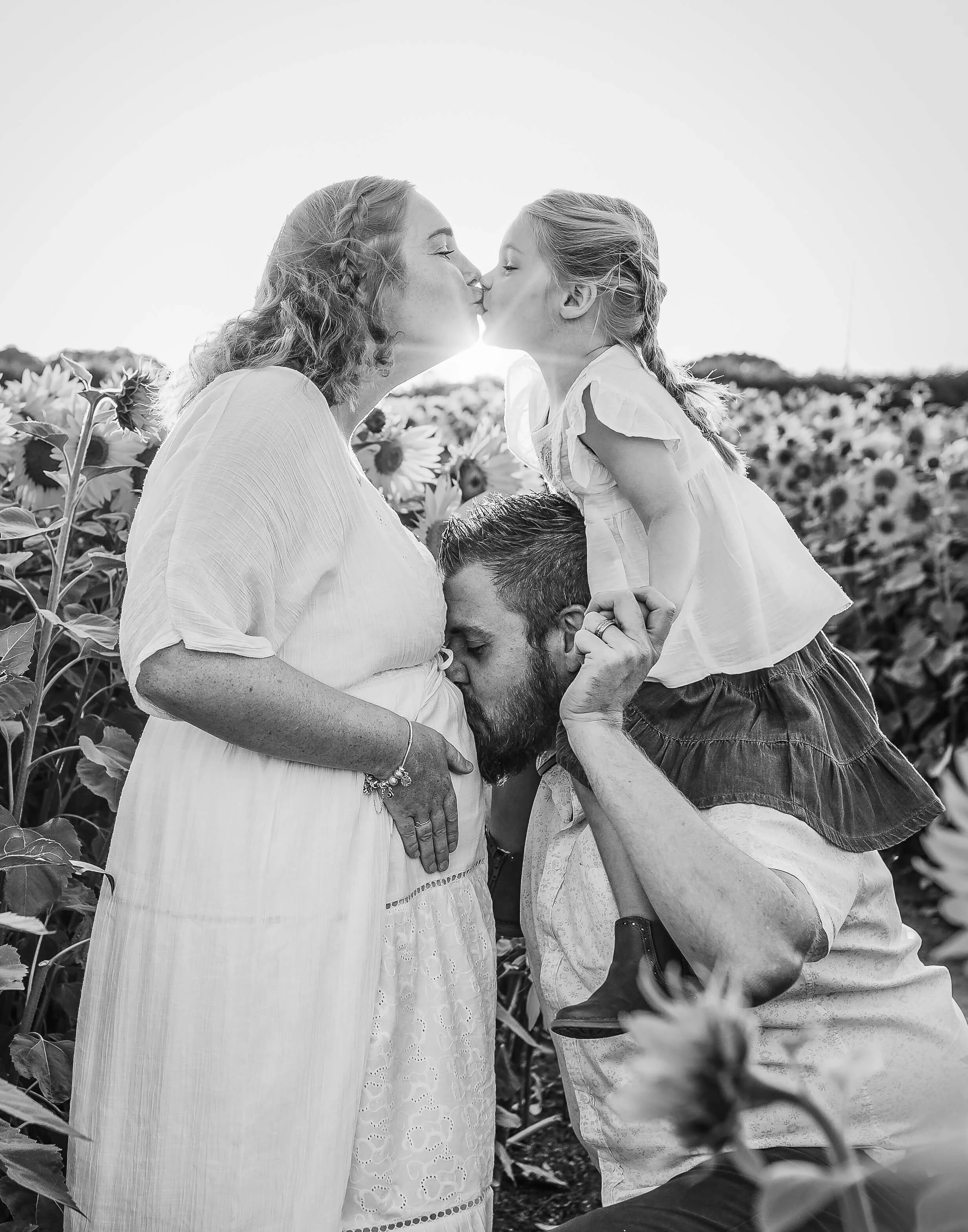 A family of three in a sunflower field, where the pregnant mother is kissing her daughter, and the father is kissing the mother's belly.