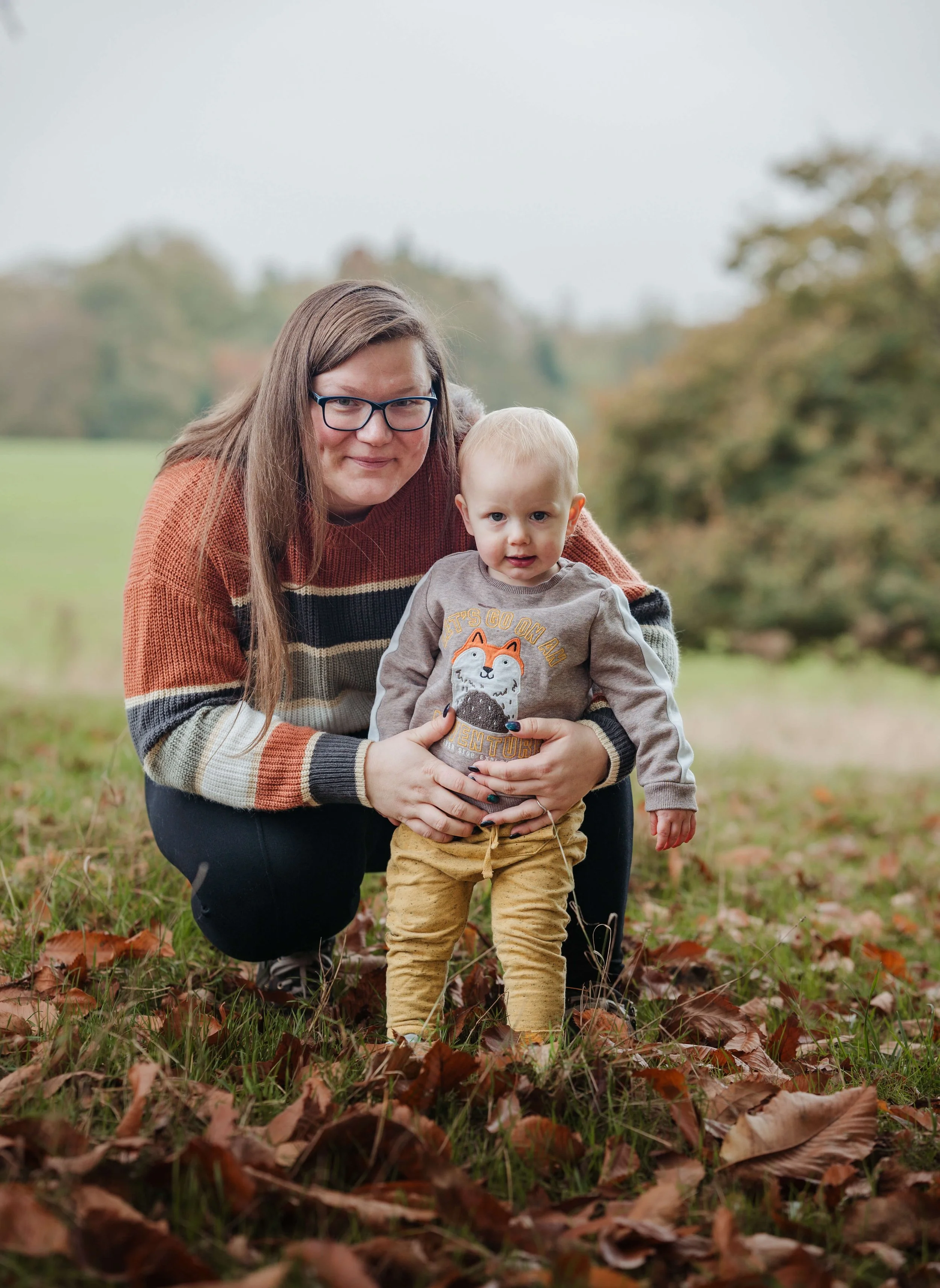 A woman with glasses wearing a striped sweater crouches down and holds a young boy with blond hair, wearing a gray sweatshirt with a fox graphic and yellow pants, in an outdoor park setting with fallen autumn leaves and trees in the background.