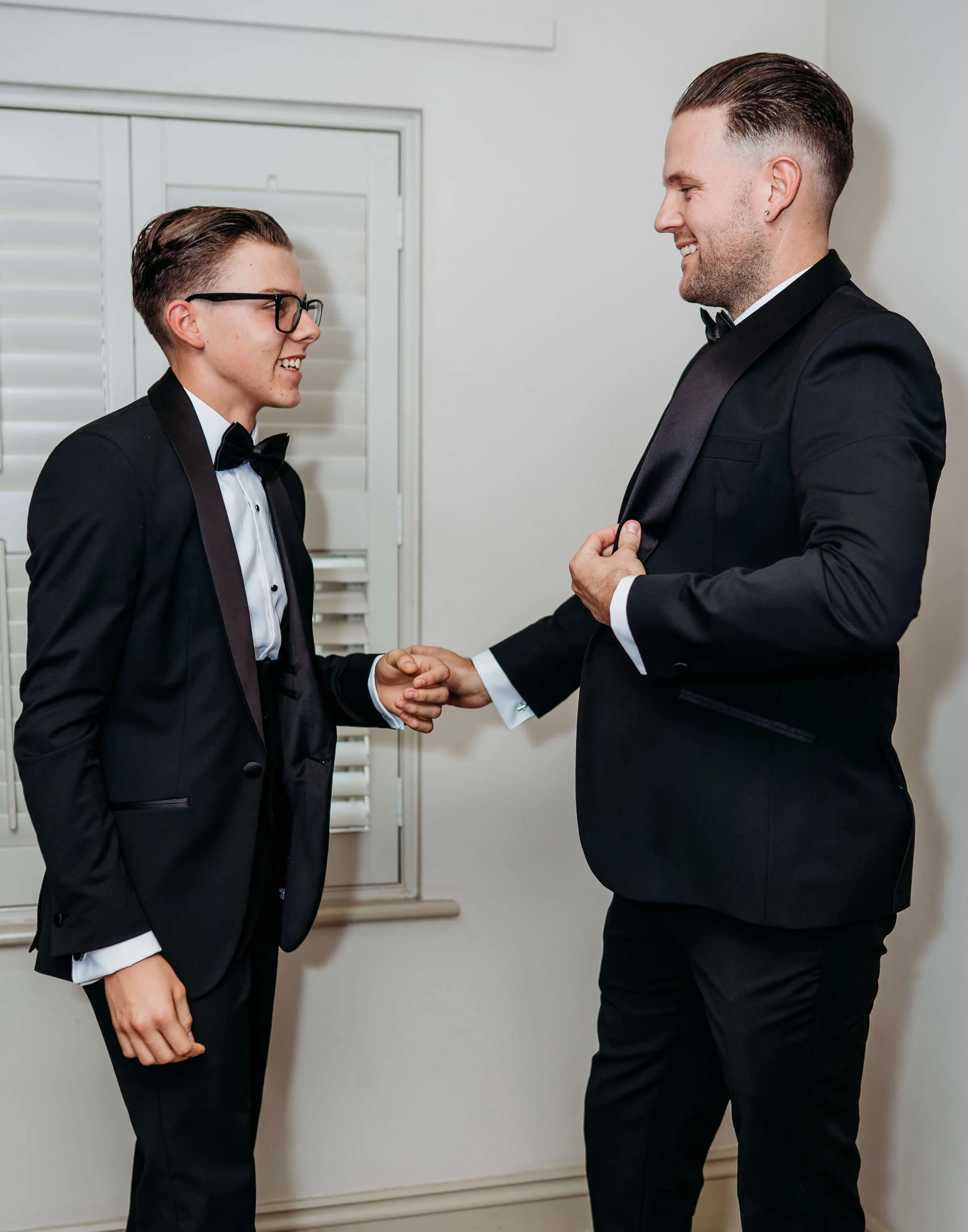 Two men in tuxedos shaking hands and smiling indoors.