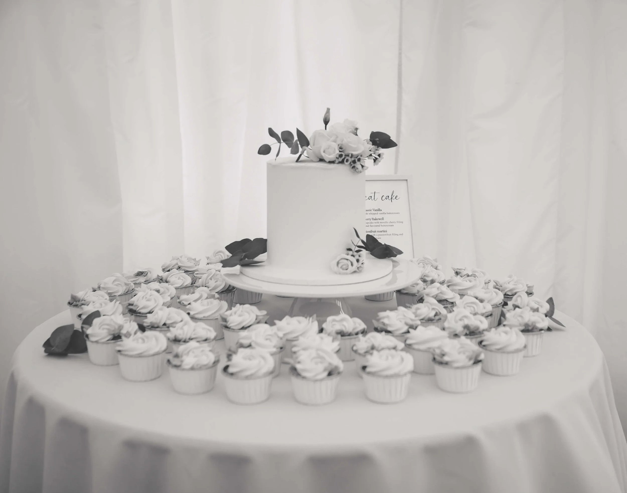 A white cake decorated with flowers on top, surrounded by many cupcakes with white frosting on a table, with a framed sign in the background.