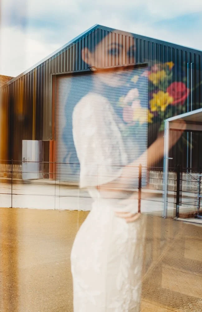 A woman holding a bouquet of colorful flowers is seen through a glass, with her reflection blending with the exterior of a modern building featuring a mural of her face.