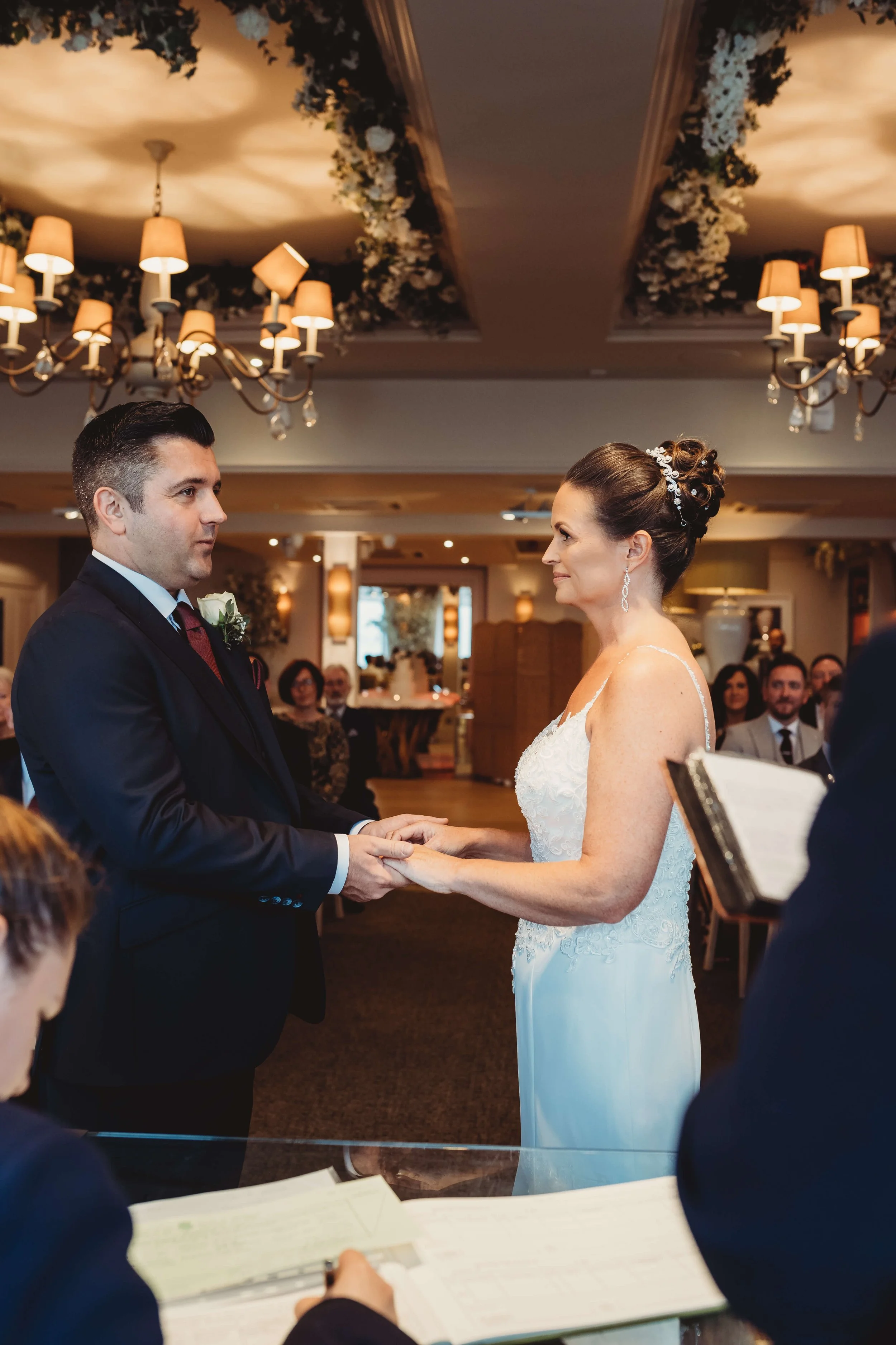 A bride and groom exchanging vows during a wedding ceremony indoors, with guests seated in the background, chandeliers hanging from the ceiling, and a decorated ceiling with flowers and greenery.