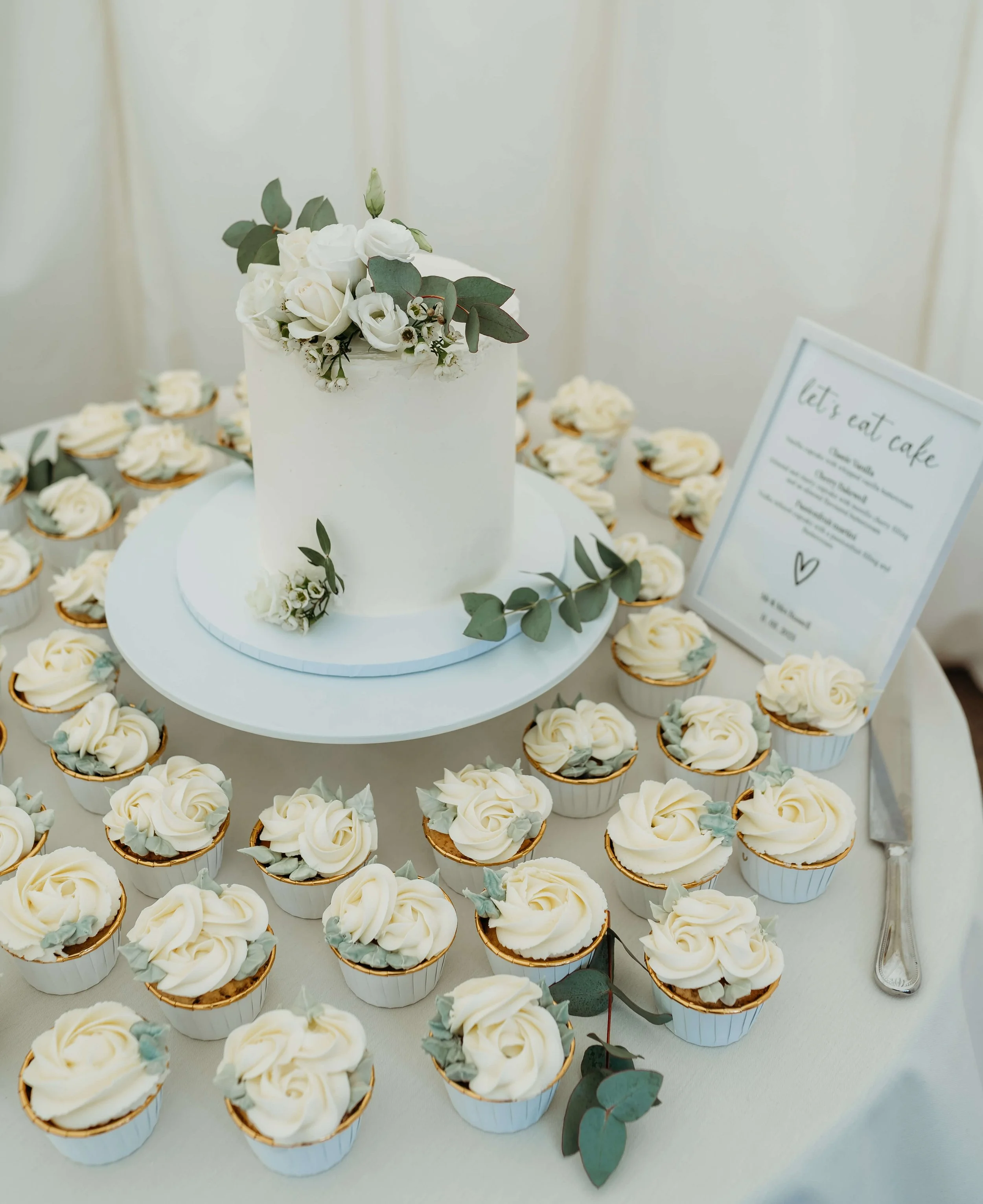 White cake with floral decoration on top surrounded by cupcakes with white frosting, some with eucalyptus leaves, on a table with a sign that says 'let's eat cake'.
