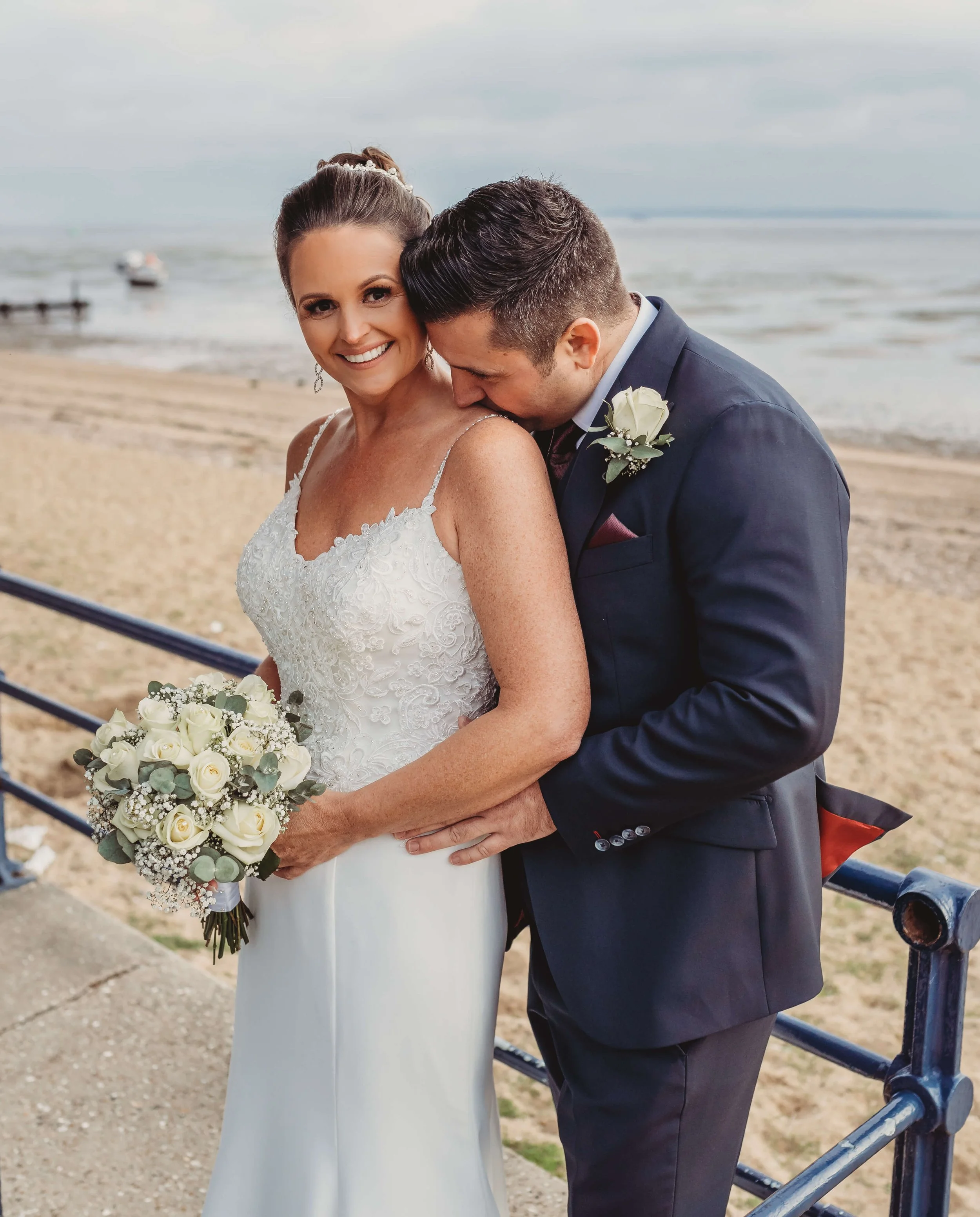 A newlywed couple standing near the beach with the groom kissing the bride's shoulder. The bride is smiling and holding a bouquet of white roses, wearing a lace wedding dress. The groom is dressed in a dark suit with a white boutonniere.