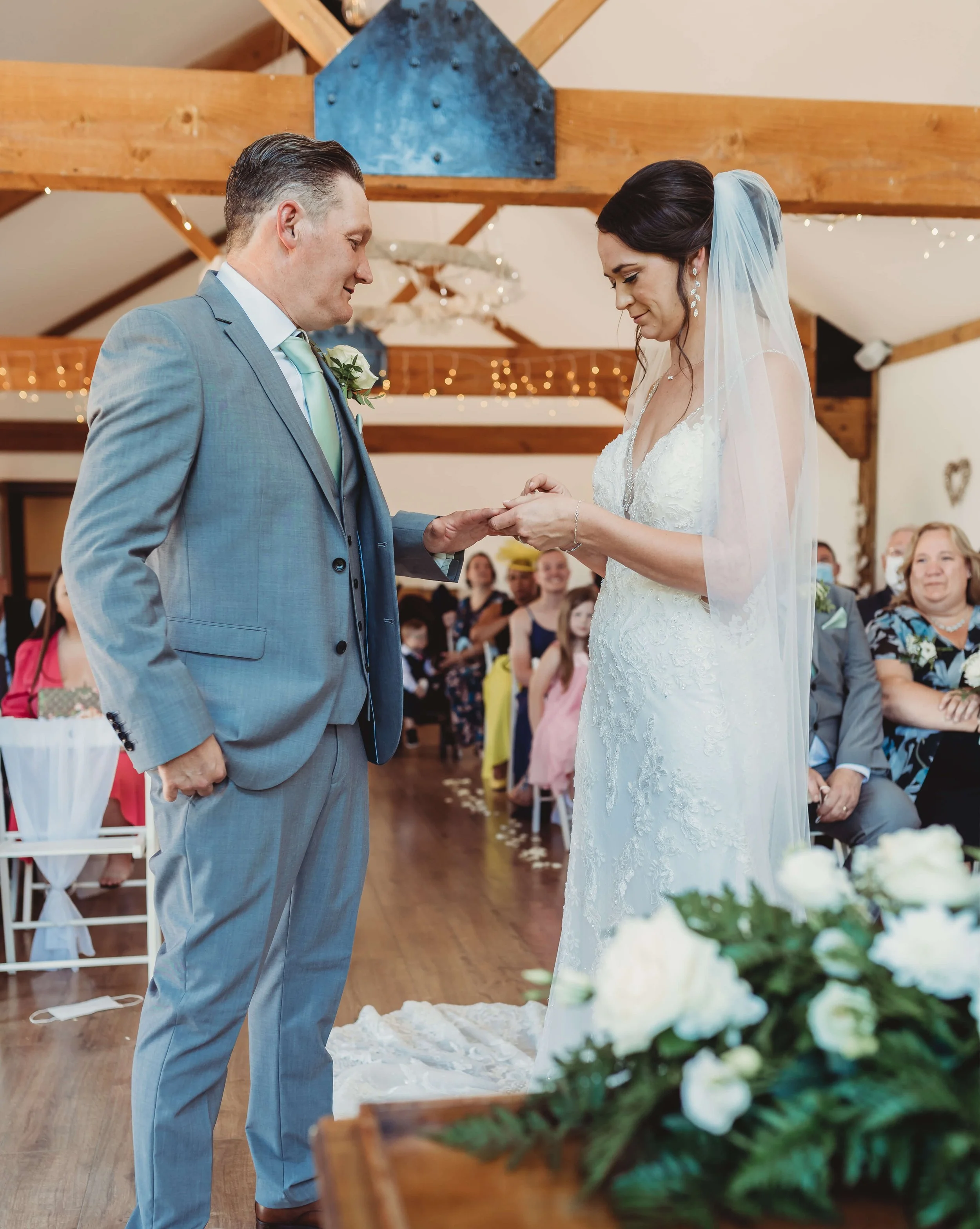 Bride and groom exchanging rings during wedding ceremony in a decorated hall, with guests seated and smiling in the background.