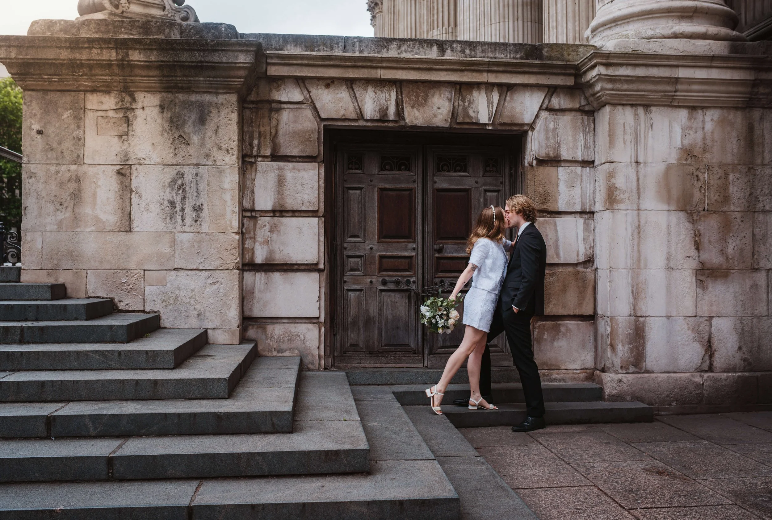 A couple dressed in wedding attire kissing on a stone staircase outside a historic building with large wooden doors.