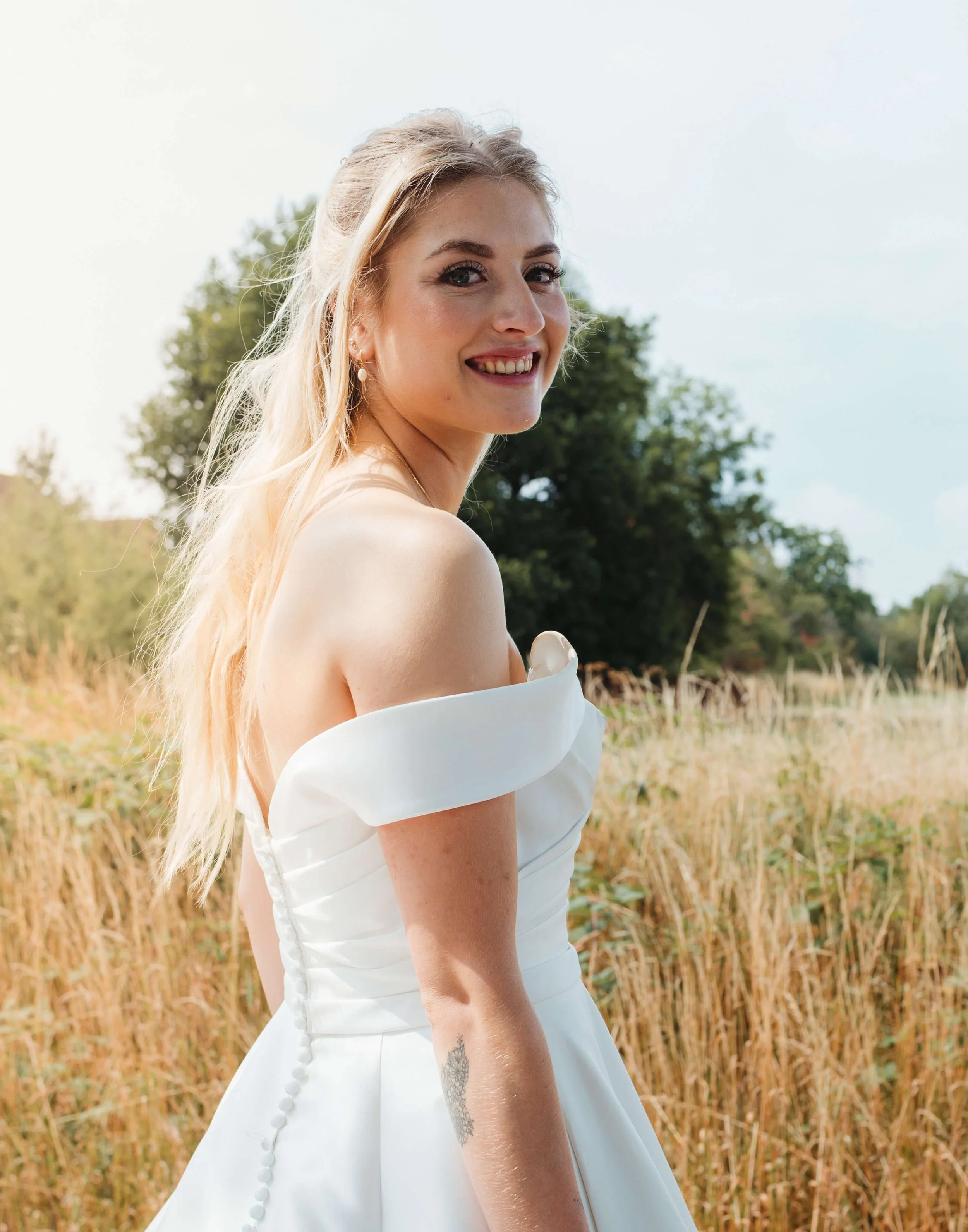 A smiling woman in a white off-shoulder dress standing outdoors in a field of tall grass with trees in the background.