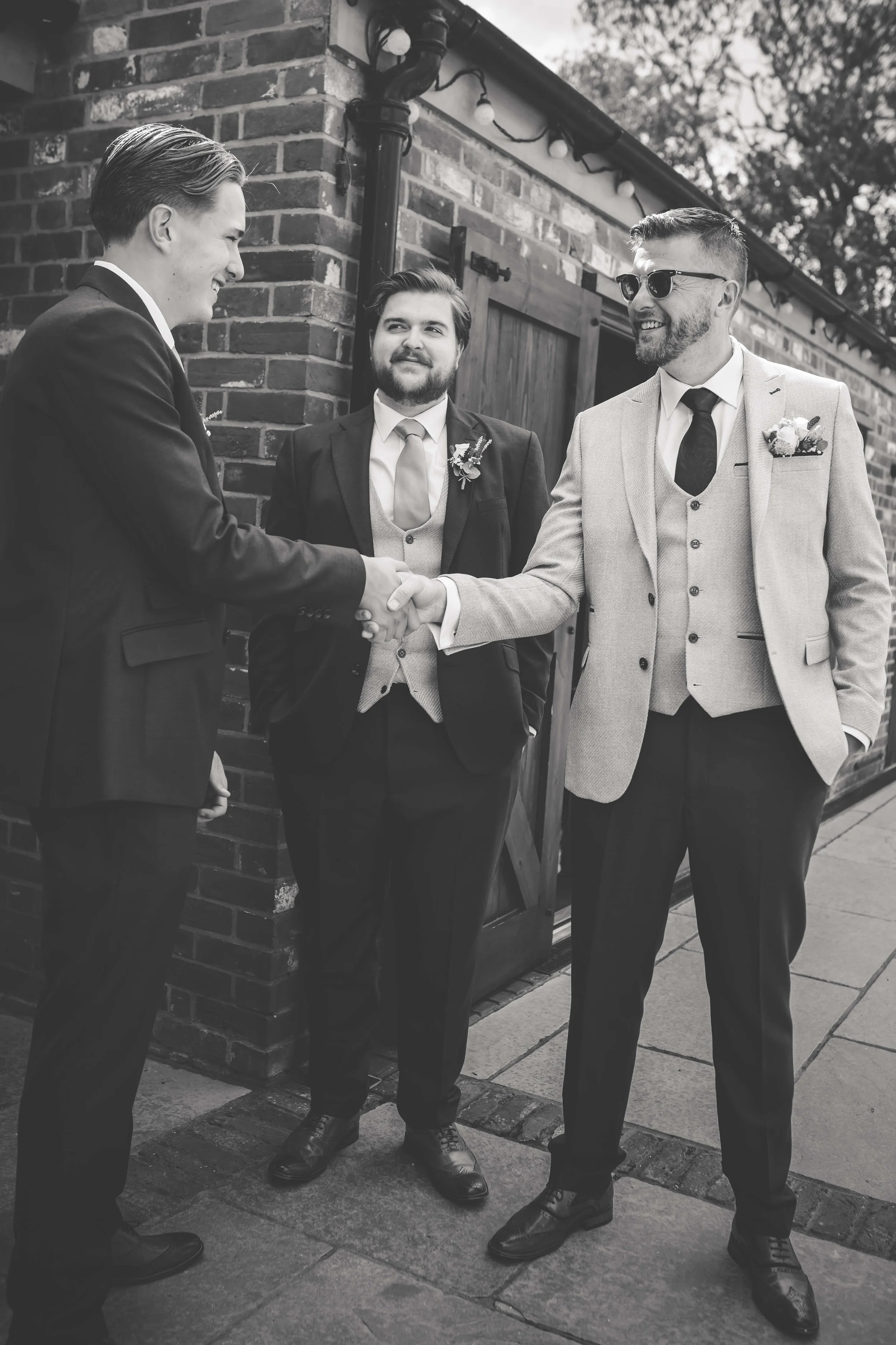Three men in suits exchanging a handshake outside a building with brick walls and wooden doors, one of them wearing sunglasses.