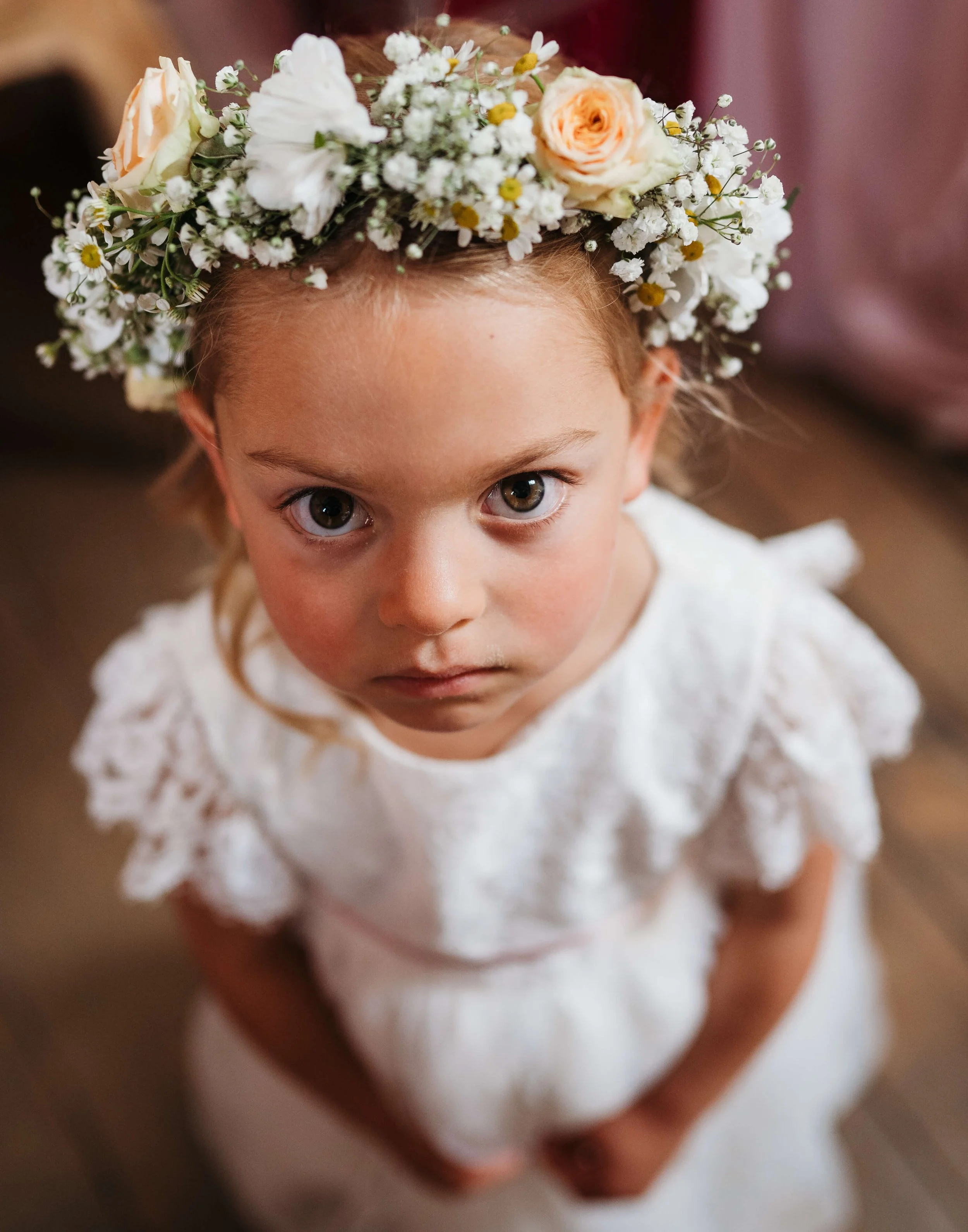 A young girl with a floral crown made of white and peach roses, baby's breath, and other small white flowers, wearing a white lace dress, looking up at the camera with wide eyes.