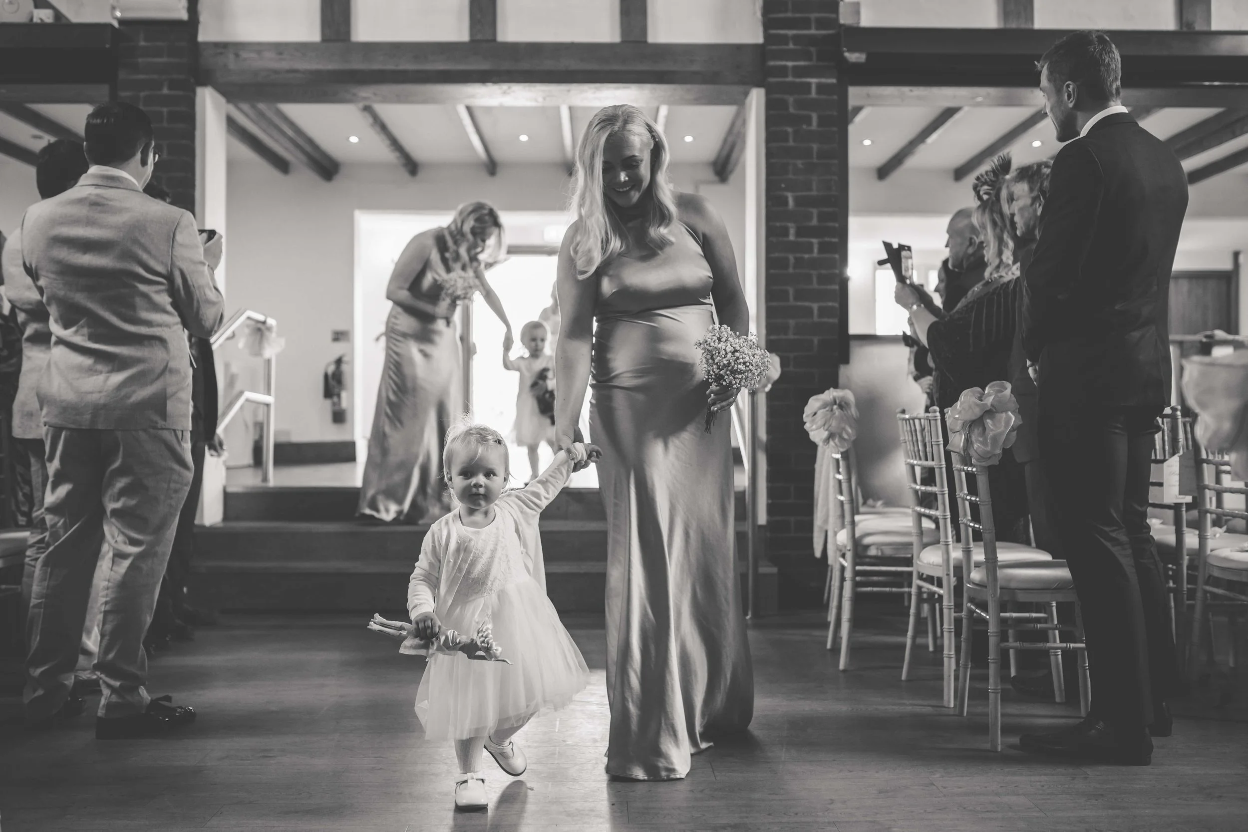 A black and white photo of a wedding scene with a woman holding a bouquet walking past a young girl holding her hand. Guests are watching and taking photos in the background, with chairs decorated with bows.