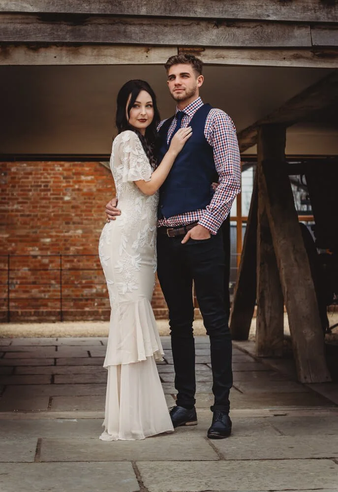 A couple dressed in formal attire, with the woman in a white lace dress and the man in a checkered shirt, vest, and dark pants, standing close together under a rustic wooden structure with a brick wall in the background.