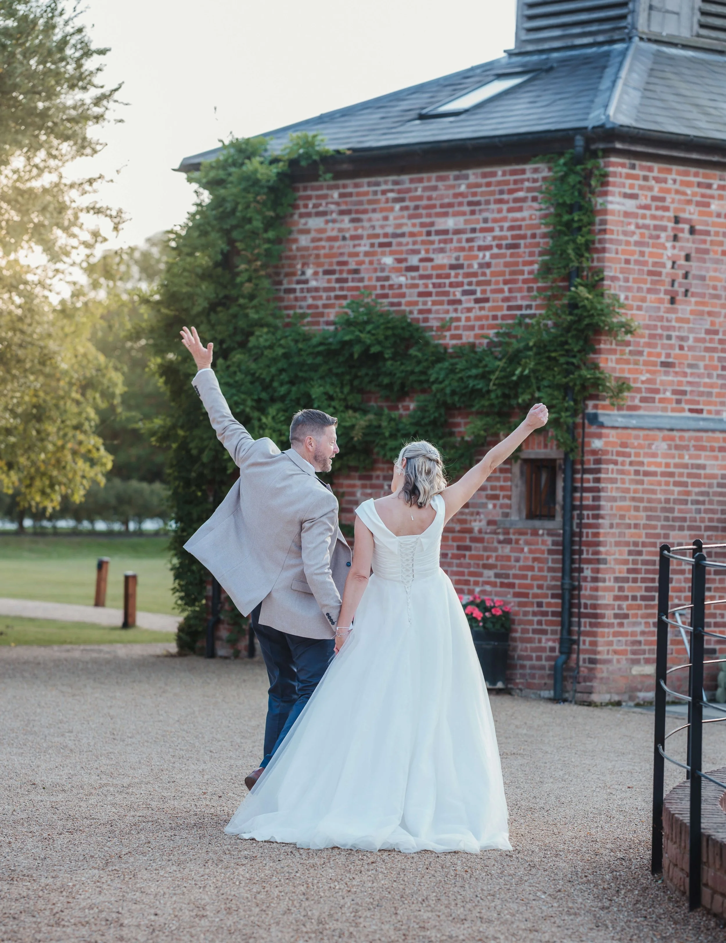 A bride and groom celebrating outdoors at sunset, holding hands and raising their arms in excitement, standing near a brick building with green vine decorations.