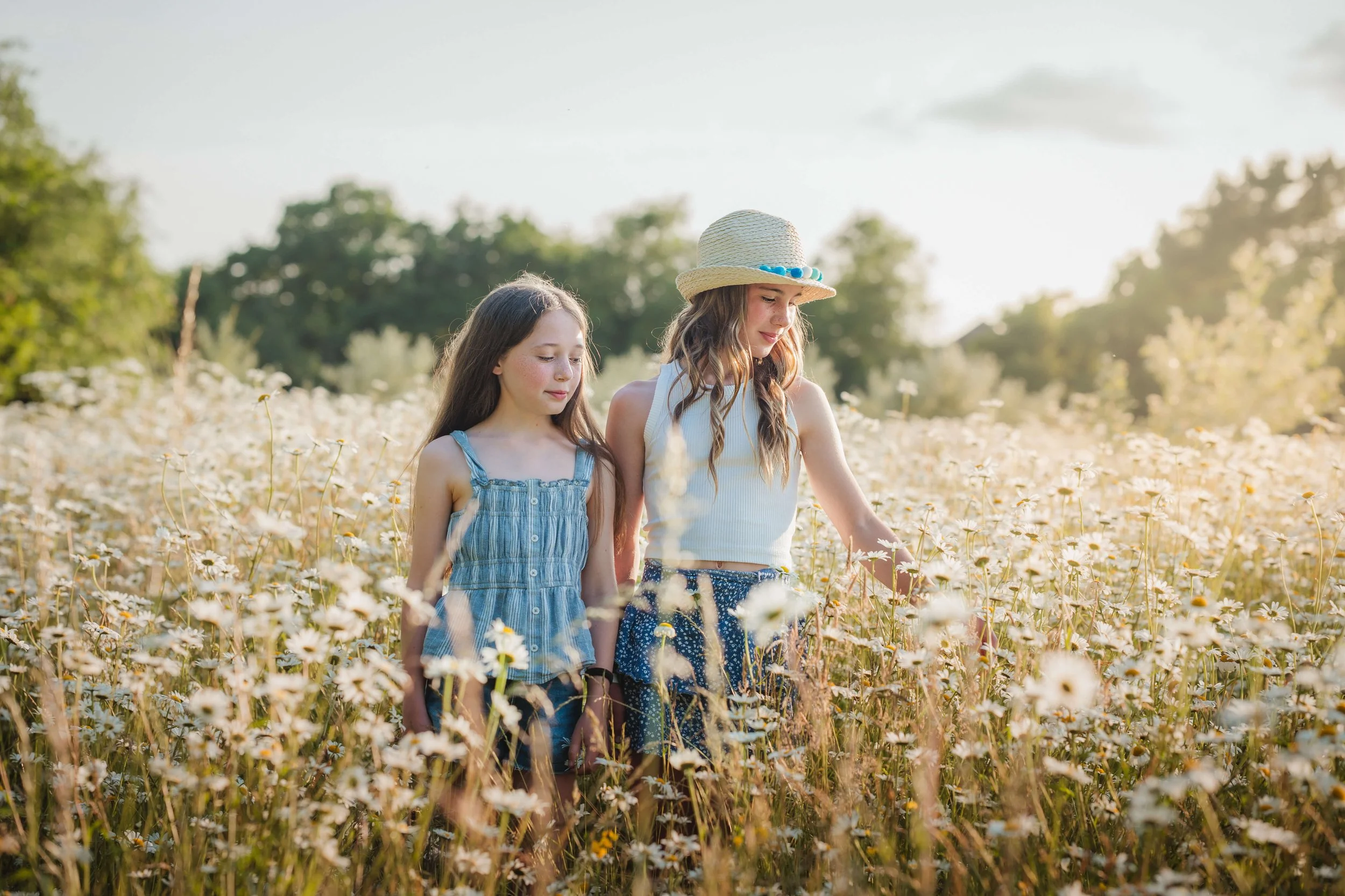 Two young girls walking through a field of white daisies, one wearing a straw hat and the other in a denim dress, during golden hour.