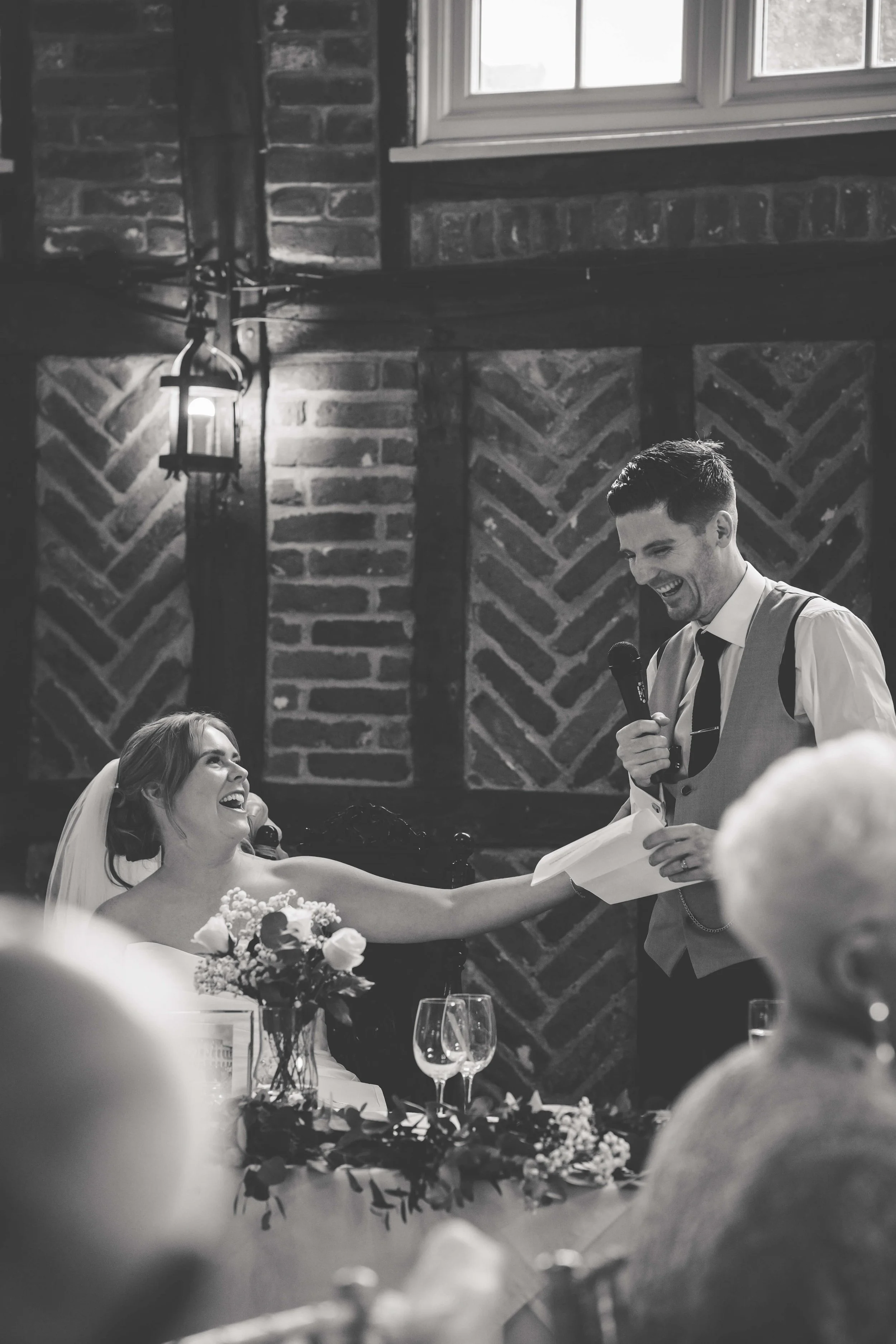 A bride and groom at their wedding reception, sharing a joyful moment with the bride reaching out to the groom who is holding a microphone and a speech paper, both smiling.