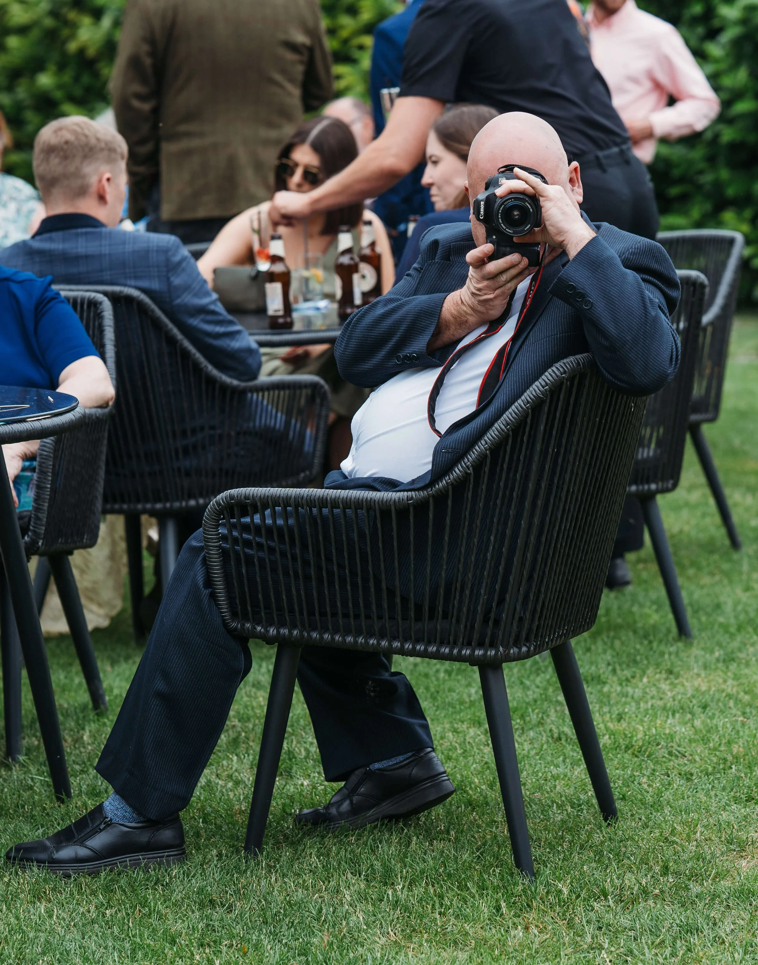 A man in a suit sitting in a black outdoor chair, holding a camera, at a social gathering or outdoor event.