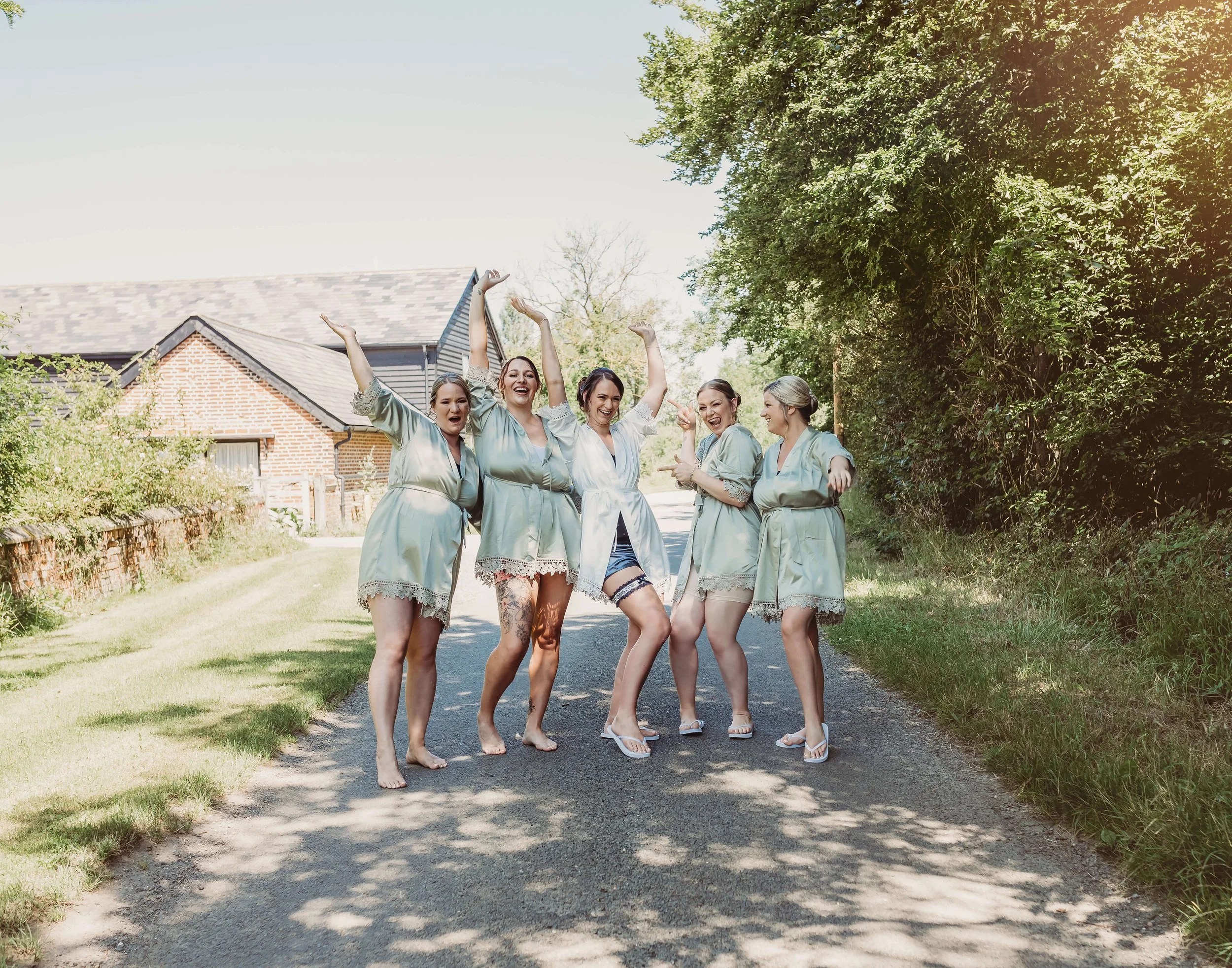 A group of five women dressed in matching silk robes and slippers, standing on a road outdoors, celebrating, smiling, and dancing with arms raised. The background features a house with a brick and siding exterior and lush green trees.