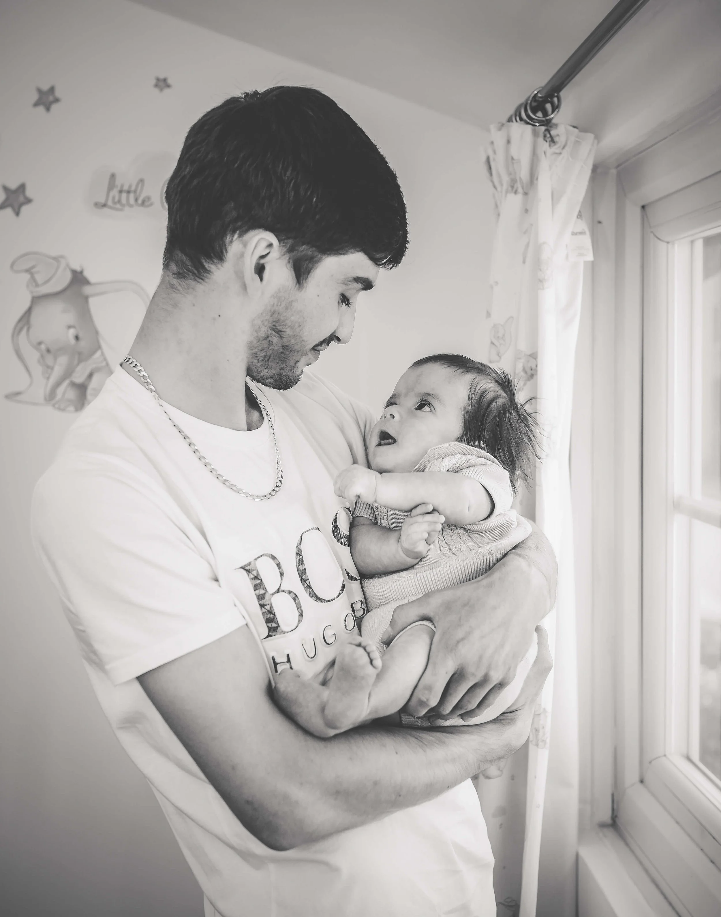A man holding a baby girl near a window, both looking at each other, in a room with cartoon character wall decals.