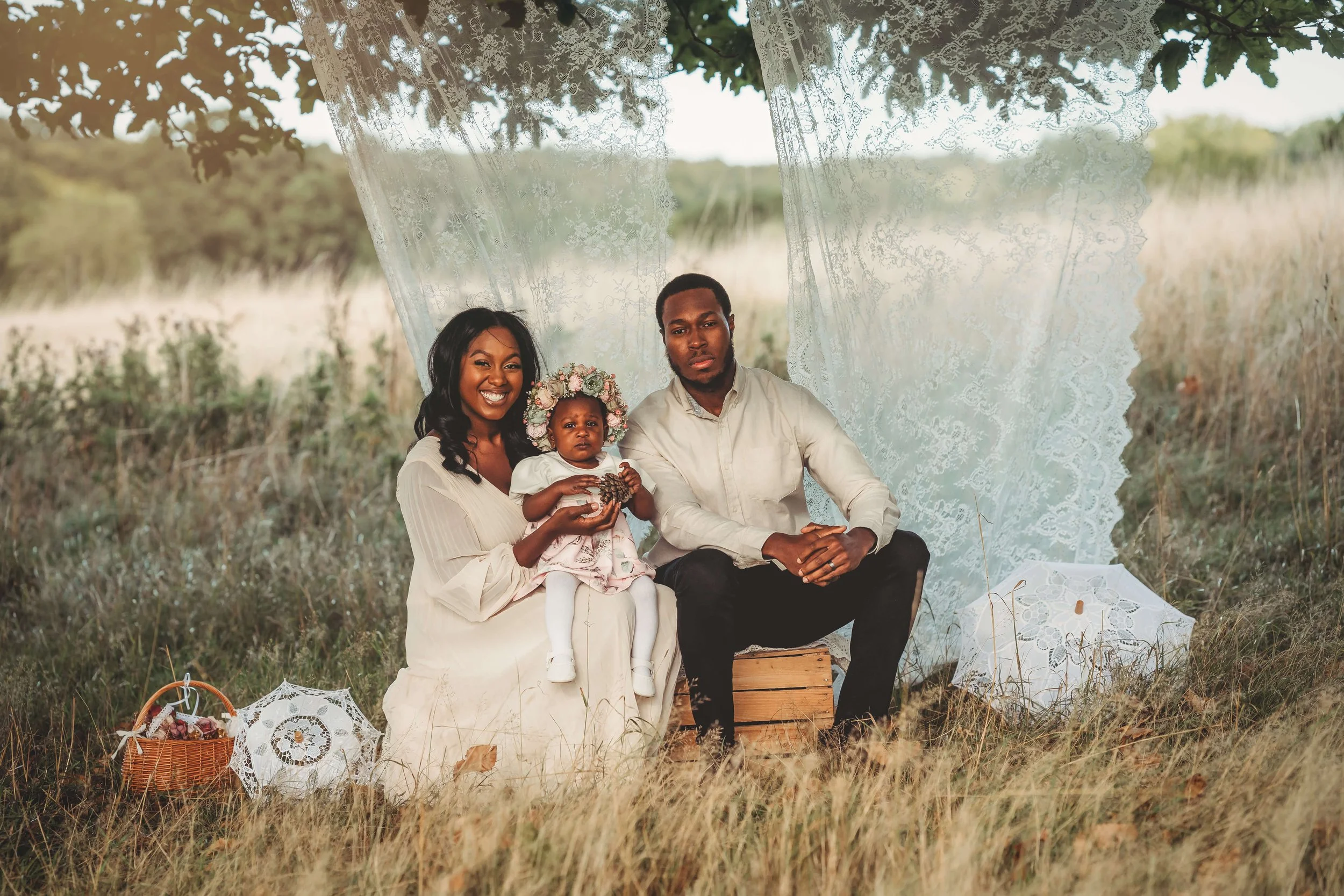 A family of three, including a woman, a man, and a young girl, sitting outdoors in a field with tall grass, under a makeshift canopy with lace curtains. The woman is holding the girl, who is wearing a floral crown, while the man sits nearby. There ar