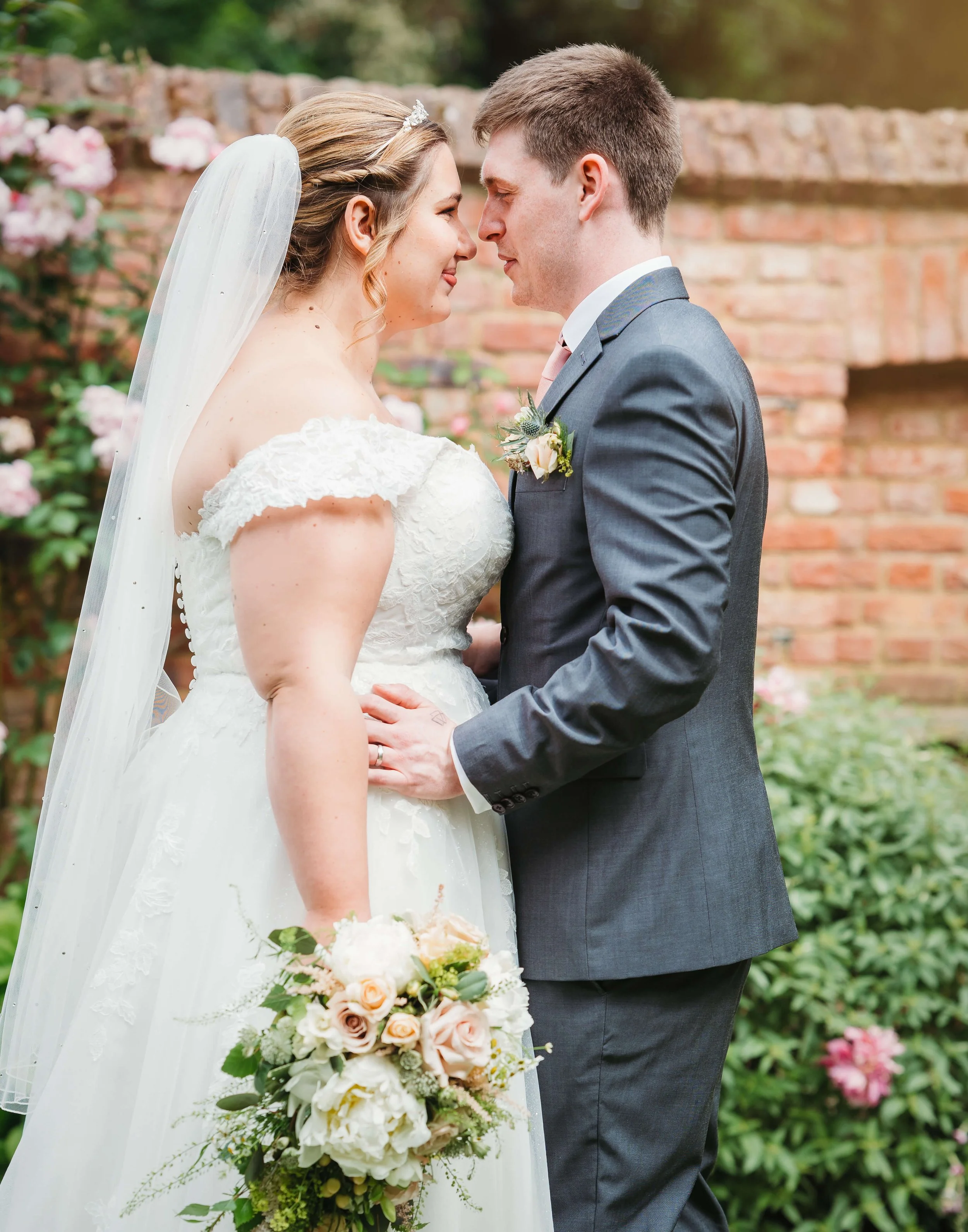 Bride and groom standing close together at their wedding, holding hands and gazing into each other's eyes outdoors surrounded by flowers and a brick wall.