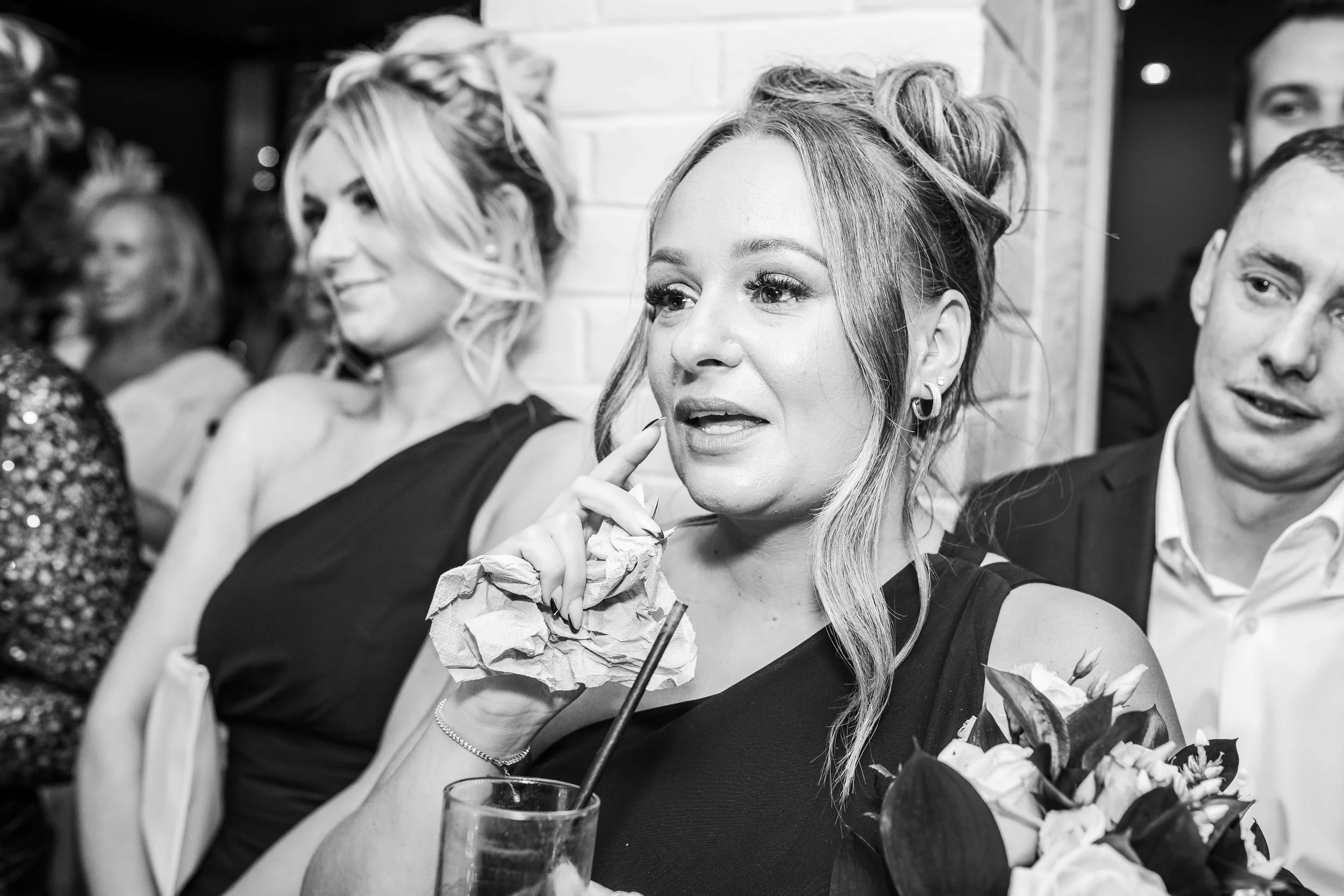 A woman with wavy hair and earrings holds a crumpled napkin and straw, appearing to be in conversation at a social event with other well-dressed people around her.