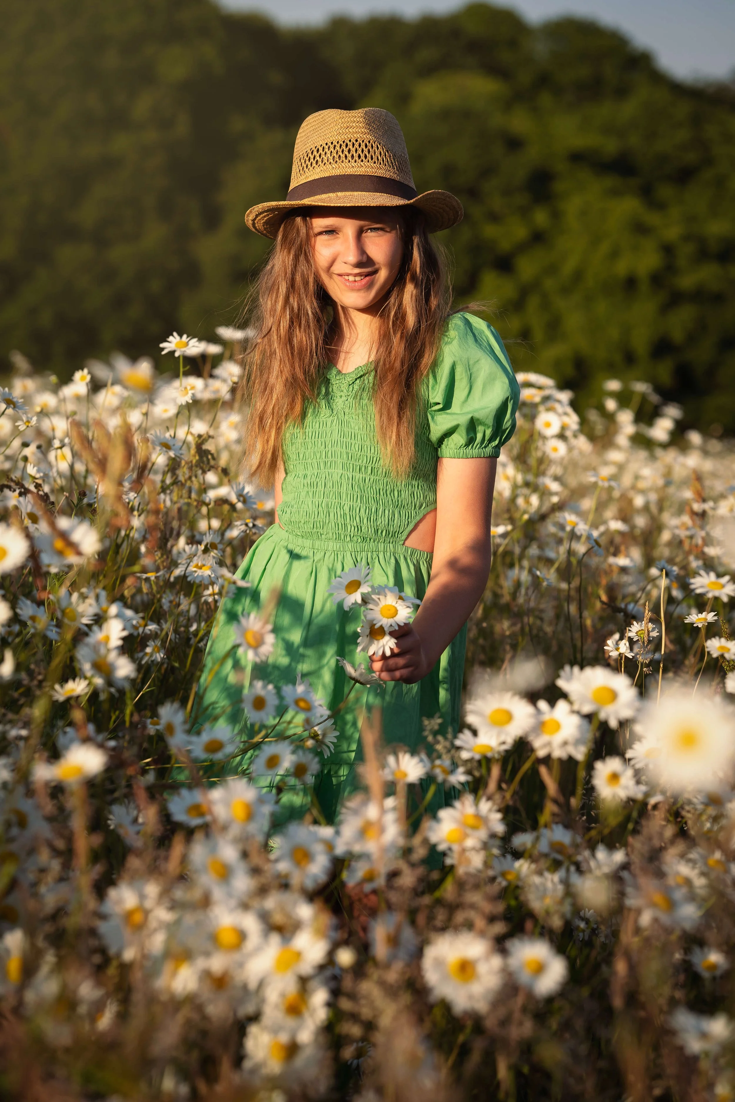 A young girl in a green dress and straw hat standing in a field of daisies during sunset.